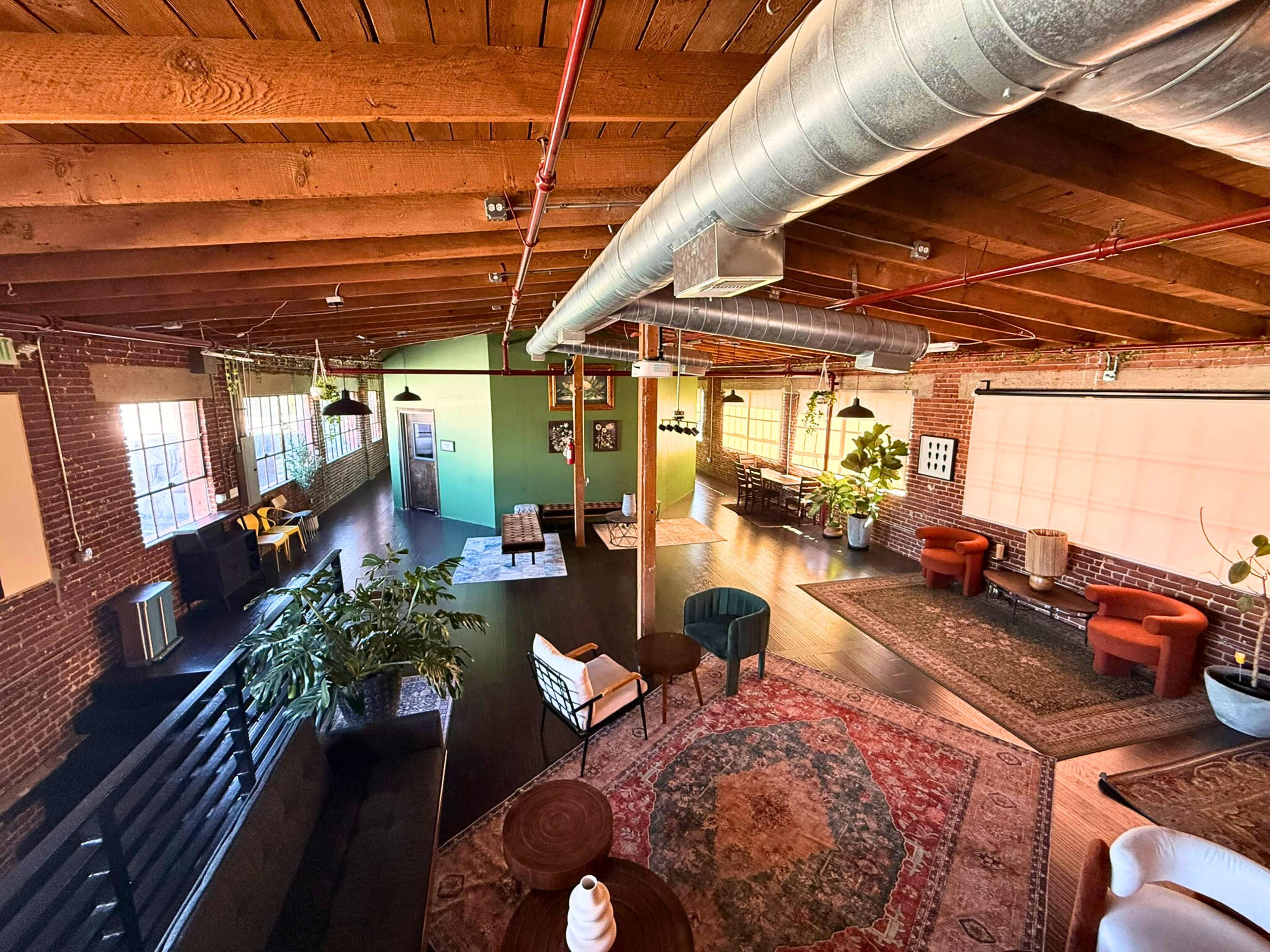 The image shows a spacious, modern office interior featuring exposed wooden beams, a mix of seating areas, and large windows that allow natural light to fill the space.