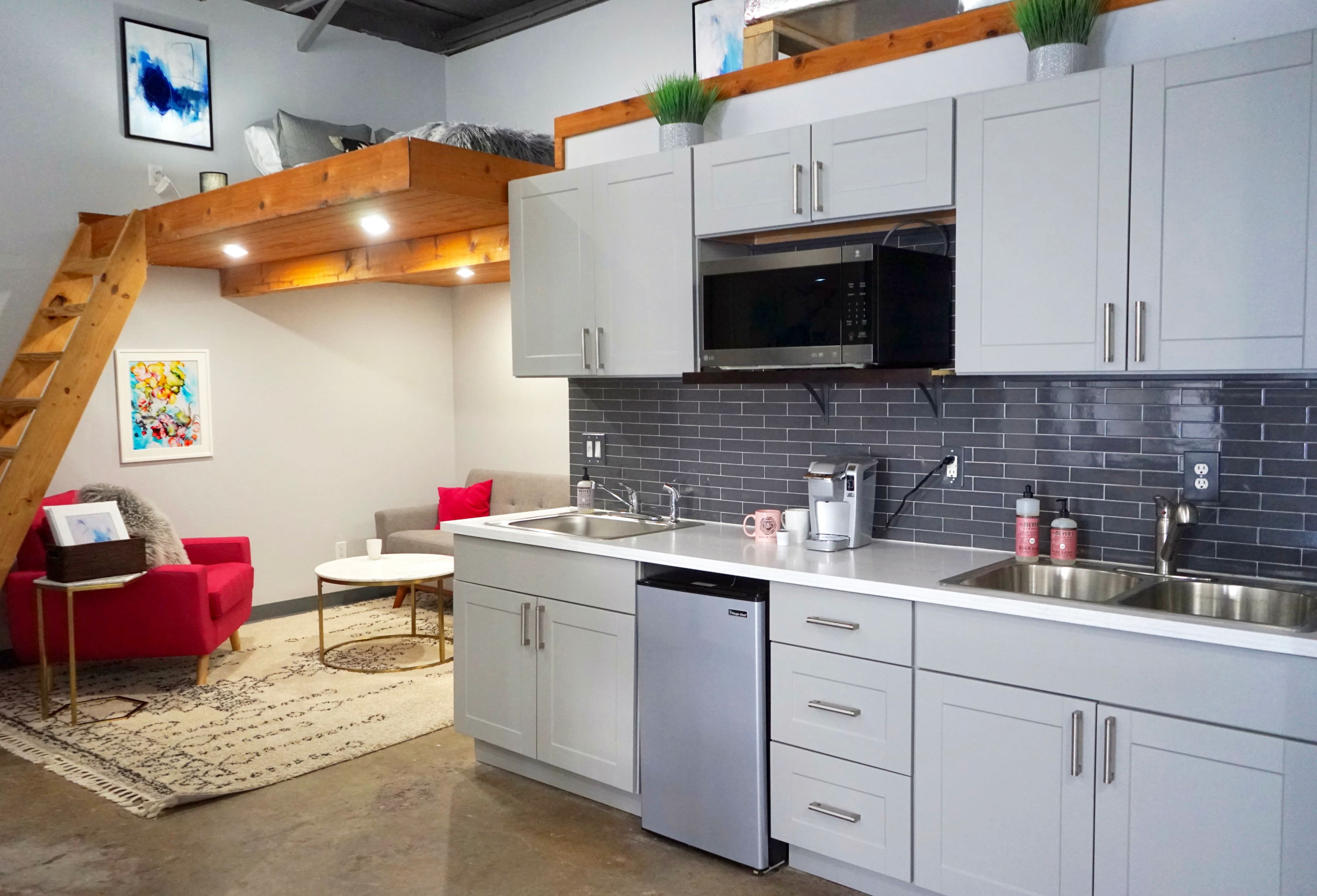 A modern kitchen area with gray cabinets, a countertop, and stainless steel appliances, alongside a cozy living space featuring a red sofa and a staircase leading to a loft.