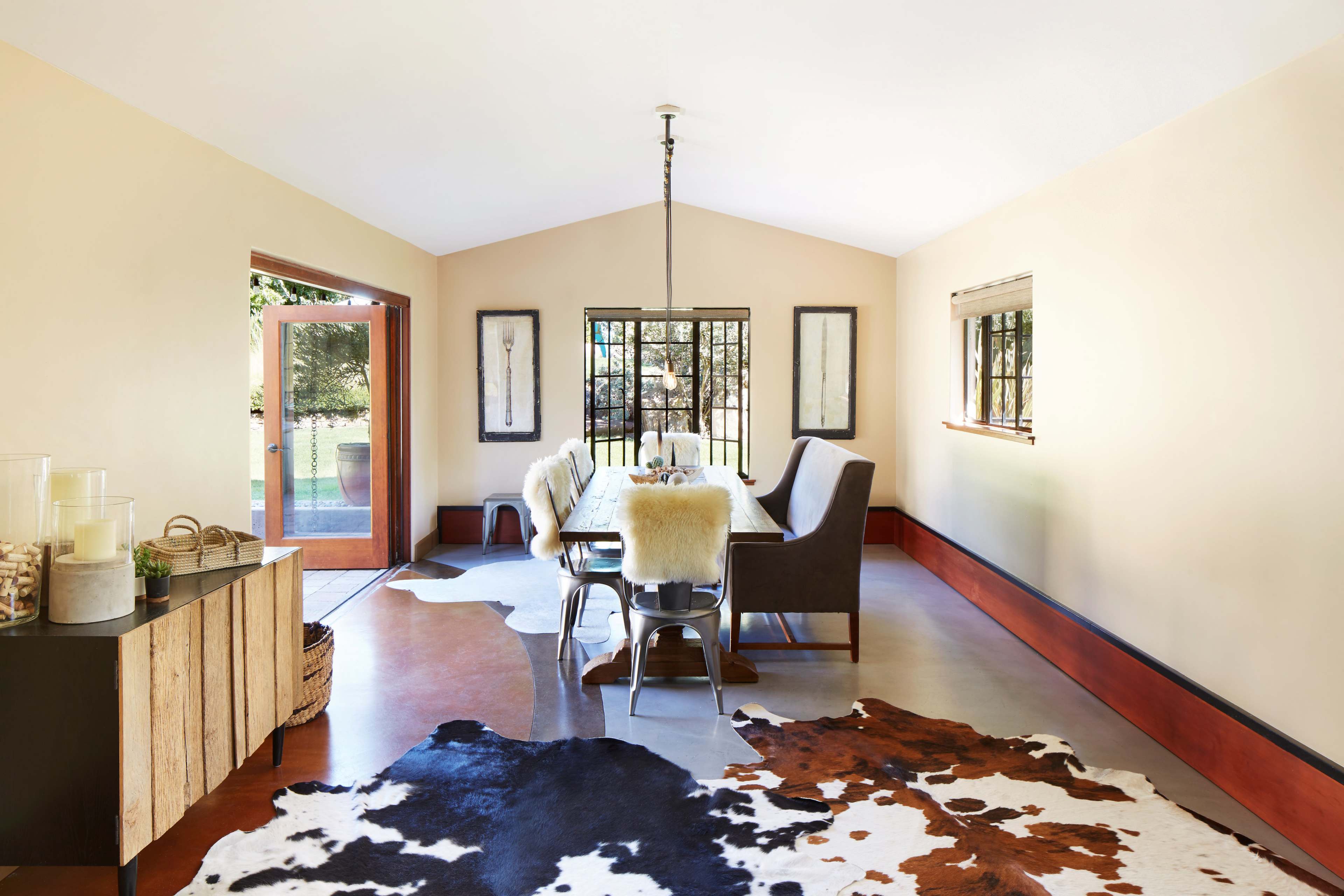The image shows a dining area with a long table surrounded by upholstered chairs, a cowhide rug on the floor, and large windows letting in natural light.
