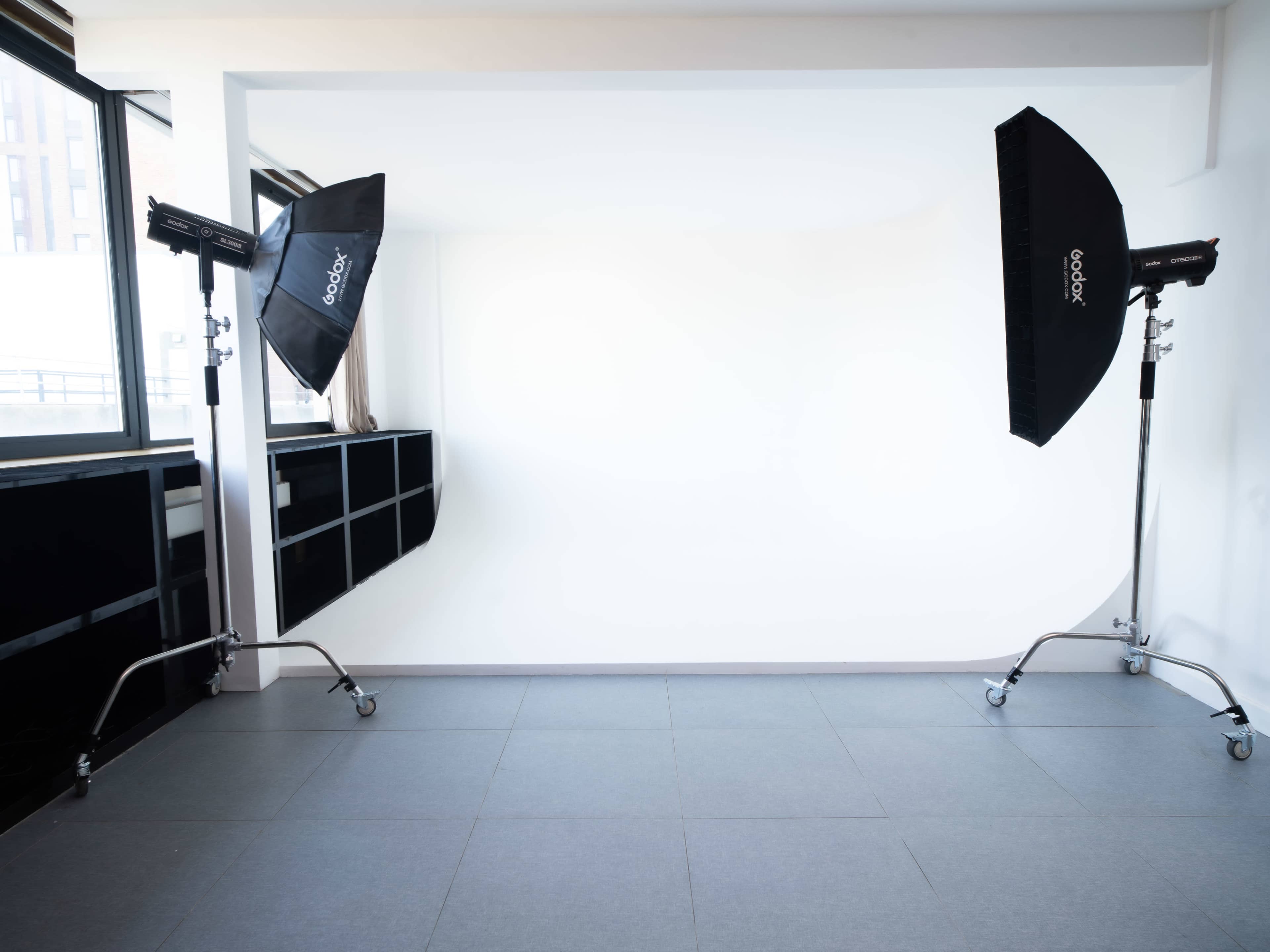 The image shows a minimalistic photography studio with two softbox lighting setups on stands positioned at either side of a blank white wall.