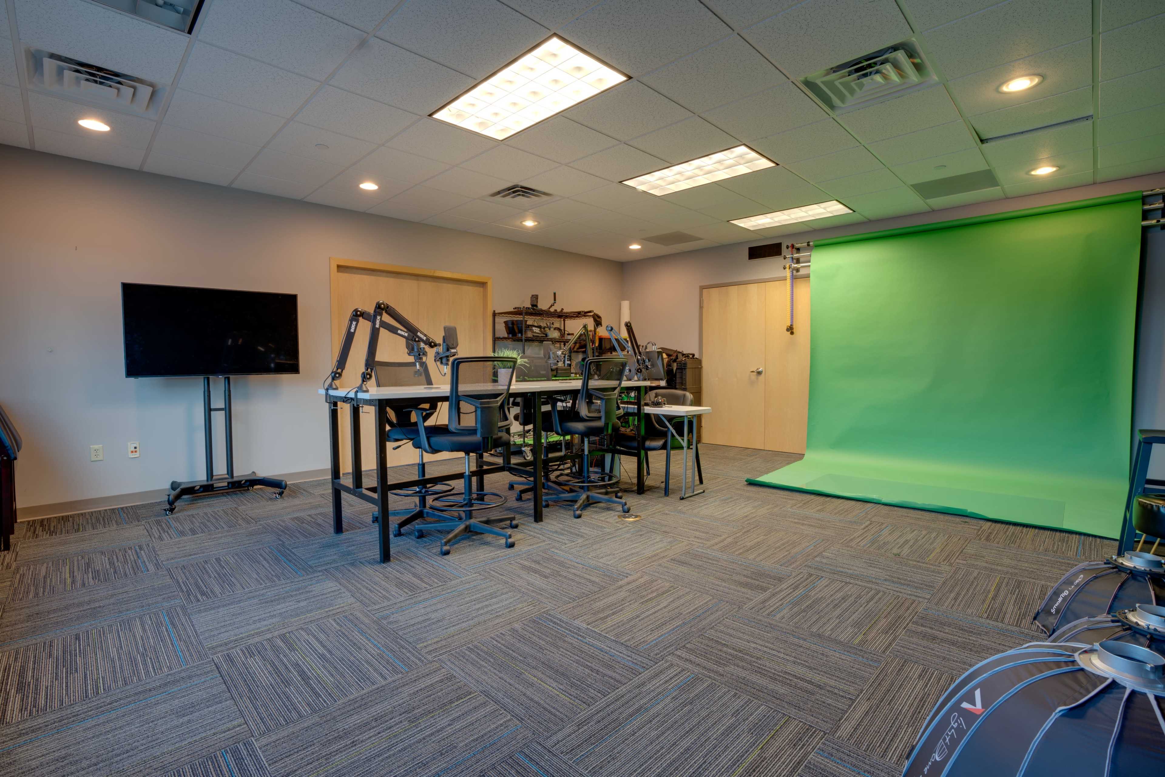 A video production studio featuring a green screen, several workstations with chairs, and a television on a stand.