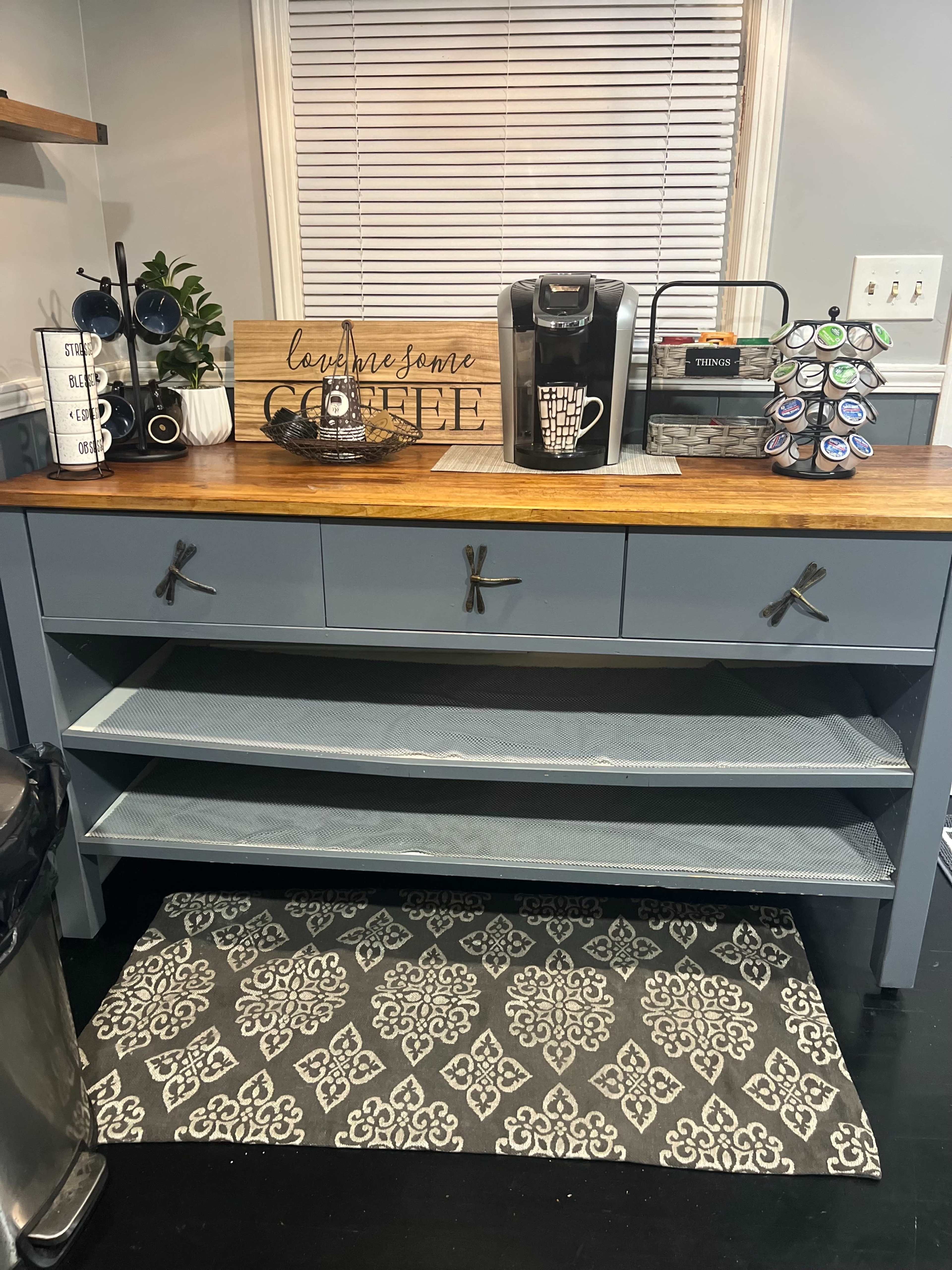 A kitchen coffee station features a wooden countertop with a coffee maker, storage drawers, and a decorative sign, set against a patterned rug.