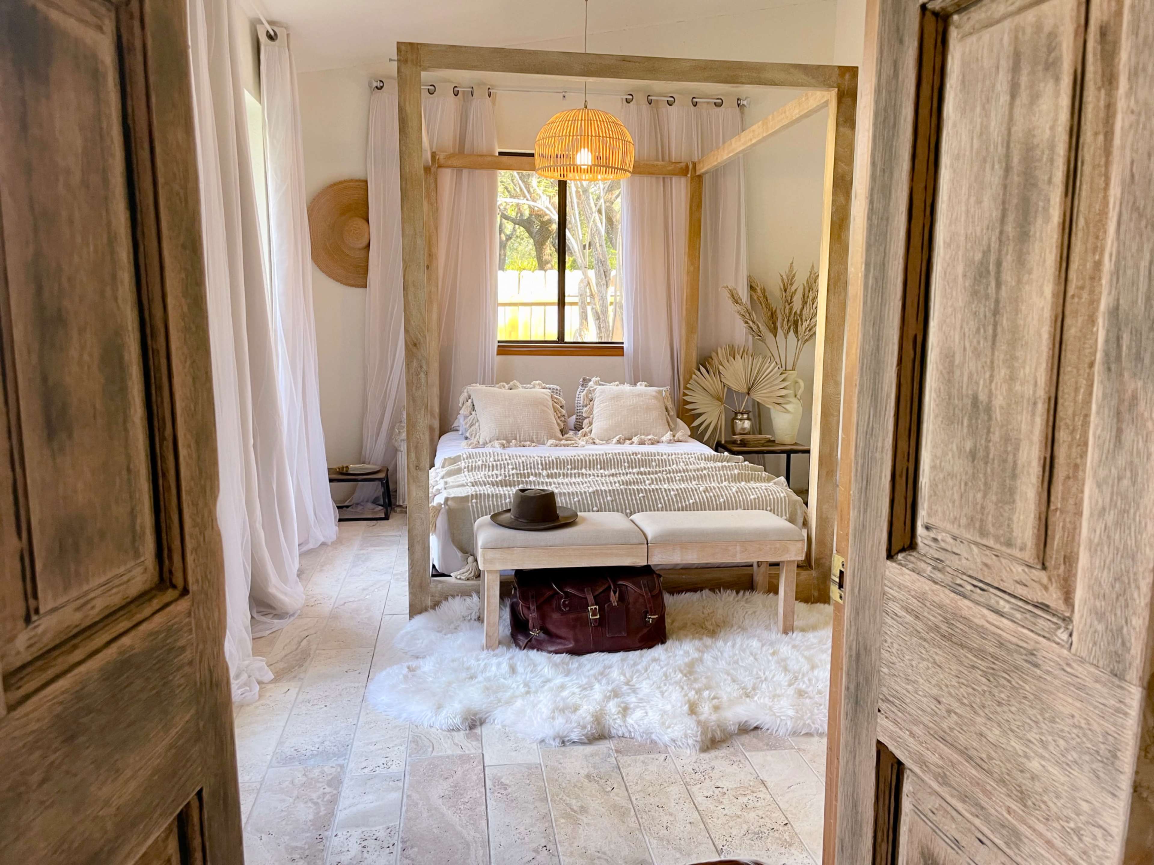 A bright bedroom features a four-poster bed with white bedding, a wooden bench, and a stylish hat placed beside a leather bag on a fluffy rug.