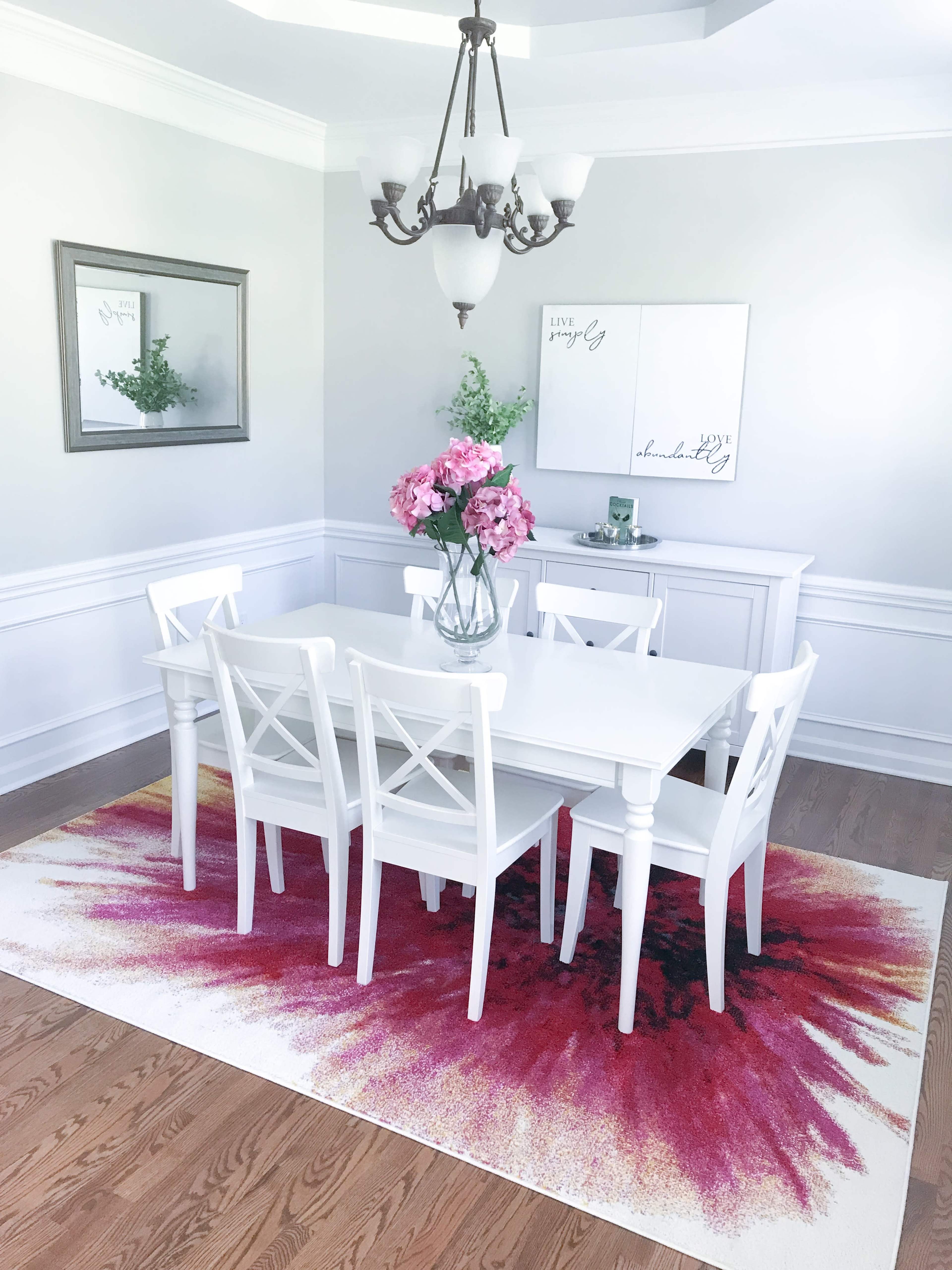 A dining area features a white table surrounded by white chairs, with a floral centerpiece and a colorful rug beneath.