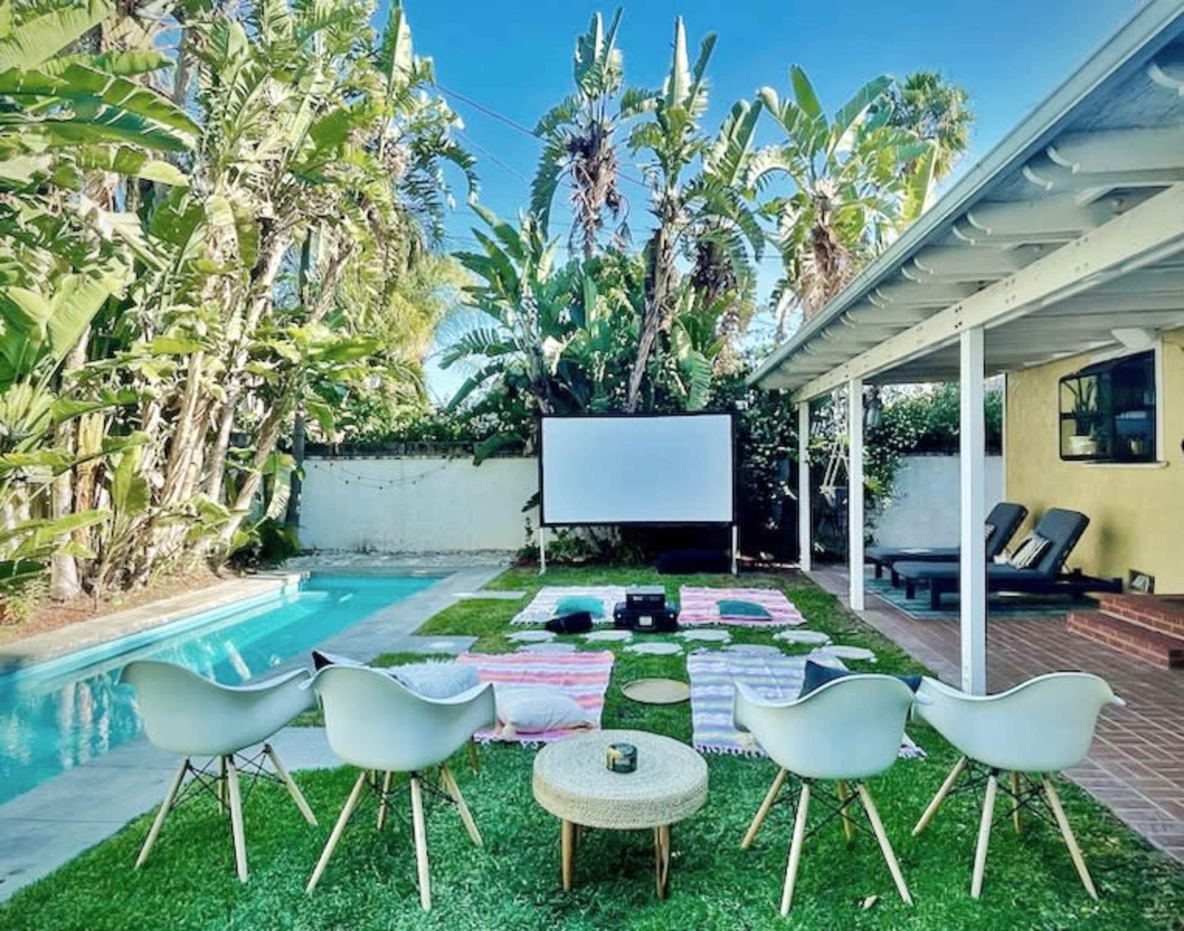 A backyard scene features a swimming pool, multiple lounge chairs, a large outdoor screen, and plush seating arranged on grassy and tiled areas, surrounded by tall palm trees.
