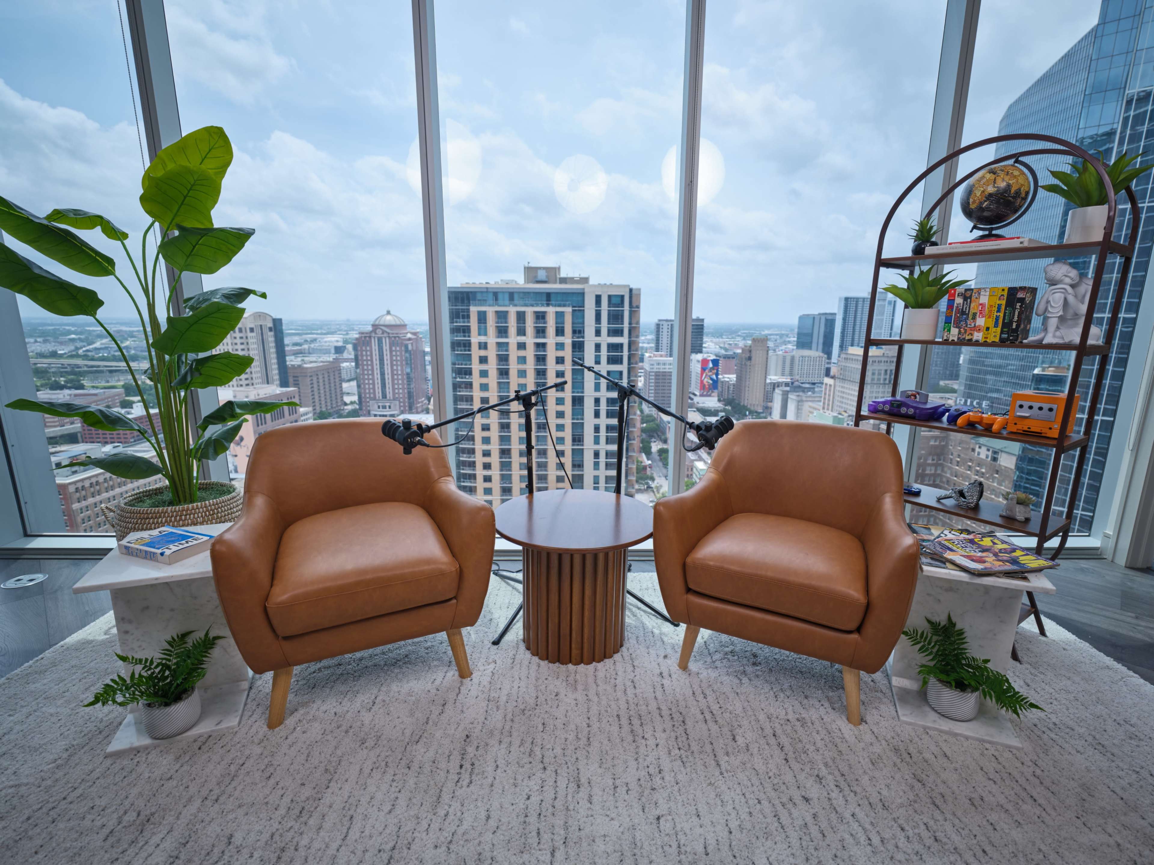 The image shows two brown chairs facing each other with a small round table in between, set in a modern high-rise room overlooking a city skyline.