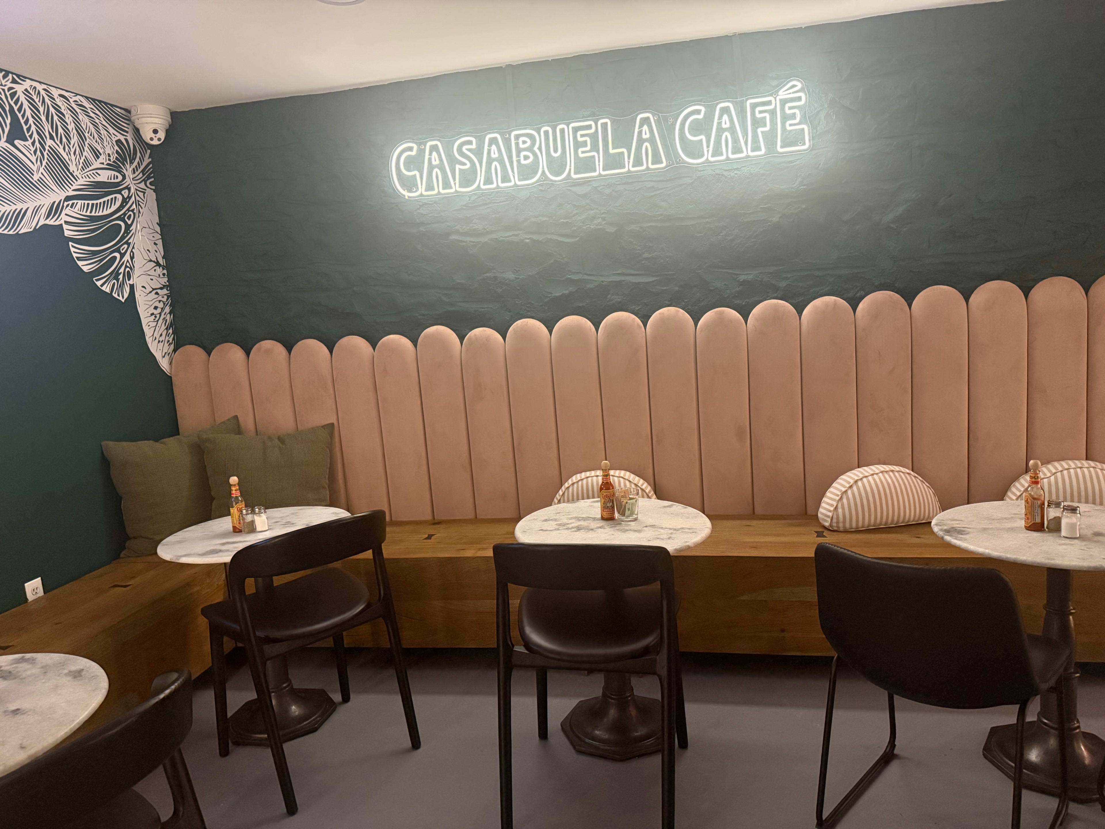 The image shows a cafe interior featuring pink upholstered wall panels, round marble tables, and dark chairs, with a neon sign that reads "CASABUELA CAFÉ."