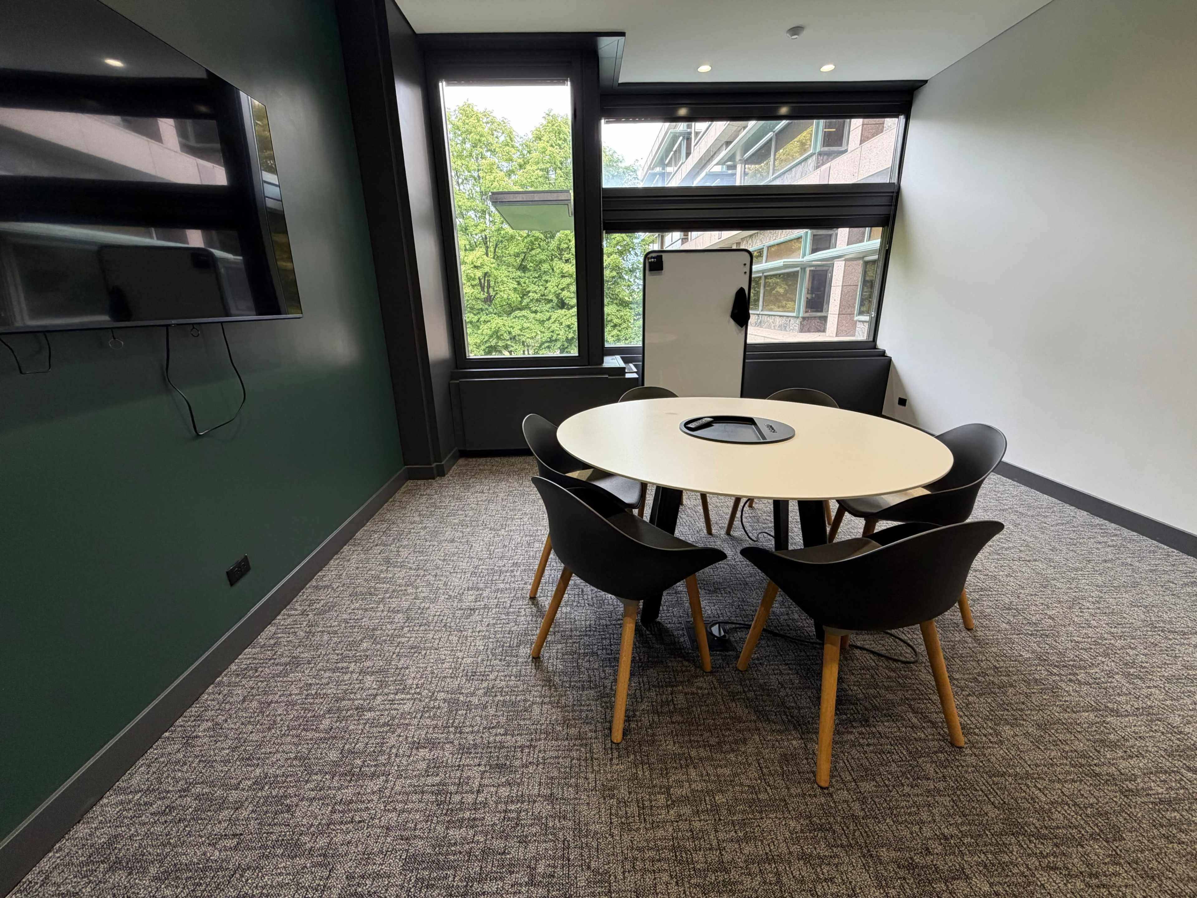 The image shows a modern conference room with a round table surrounded by four black chairs, a wall-mounted screen, and large windows with views of greenery outside.