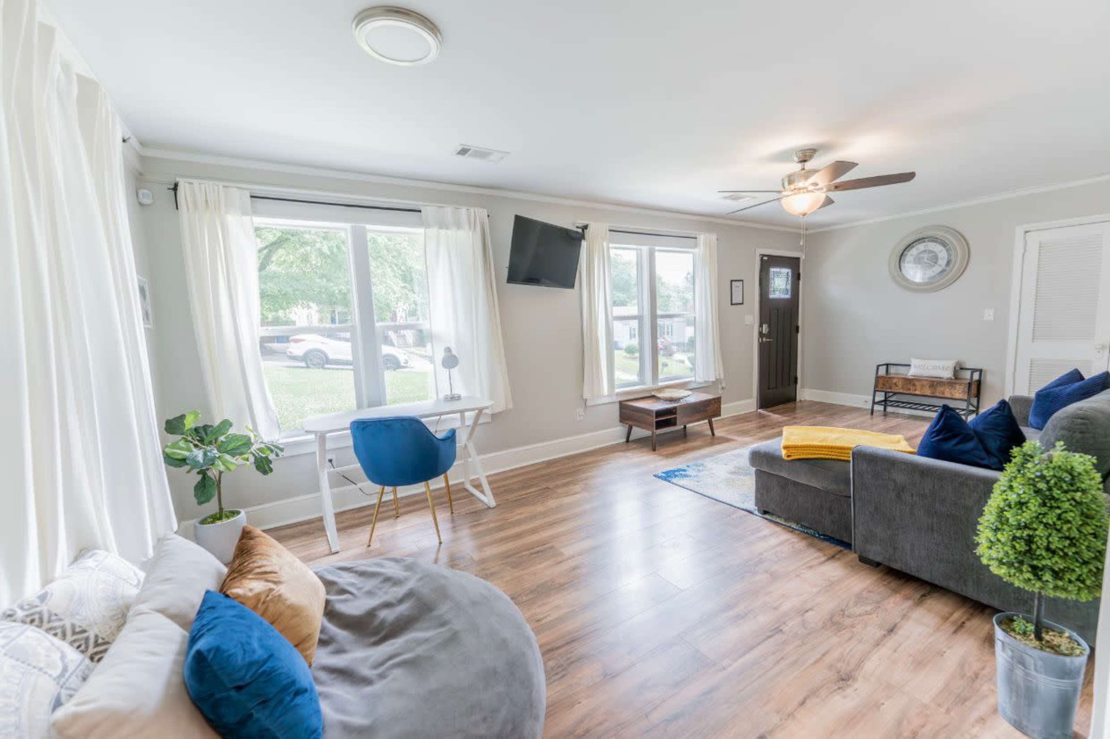 A spacious living room features a gray sofa, a small wooden coffee table, a decorative plant, and large windows allowing natural light to enter.