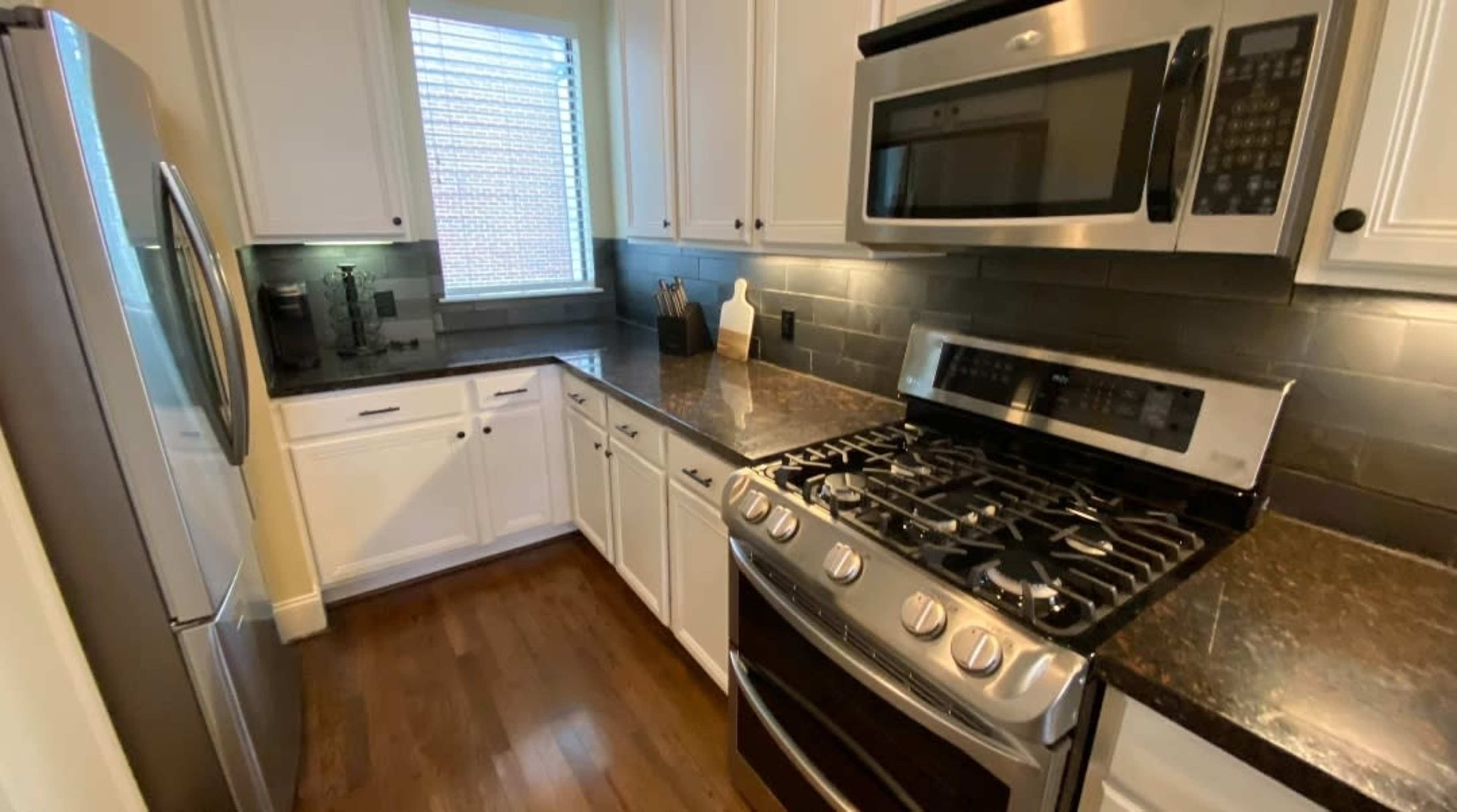 A modern kitchen with stainless steel appliances, dark countertops, white cabinets, and a window letting in natural light.