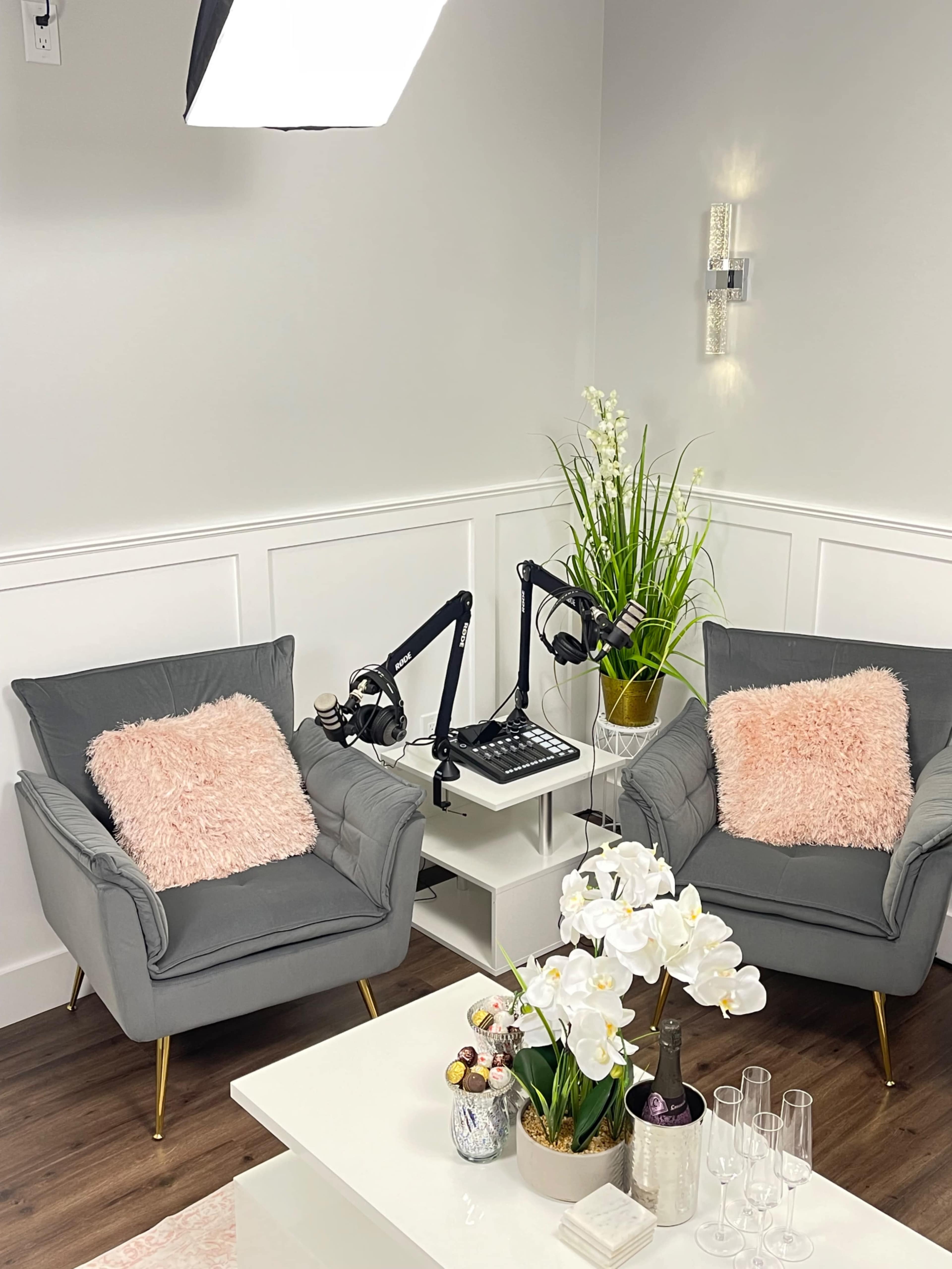 The image shows a cozy recording setup featuring two gray armchairs with pink pillows, a small white table, and a microphone on an adjustable arm, adorned with a plant and white orchids.