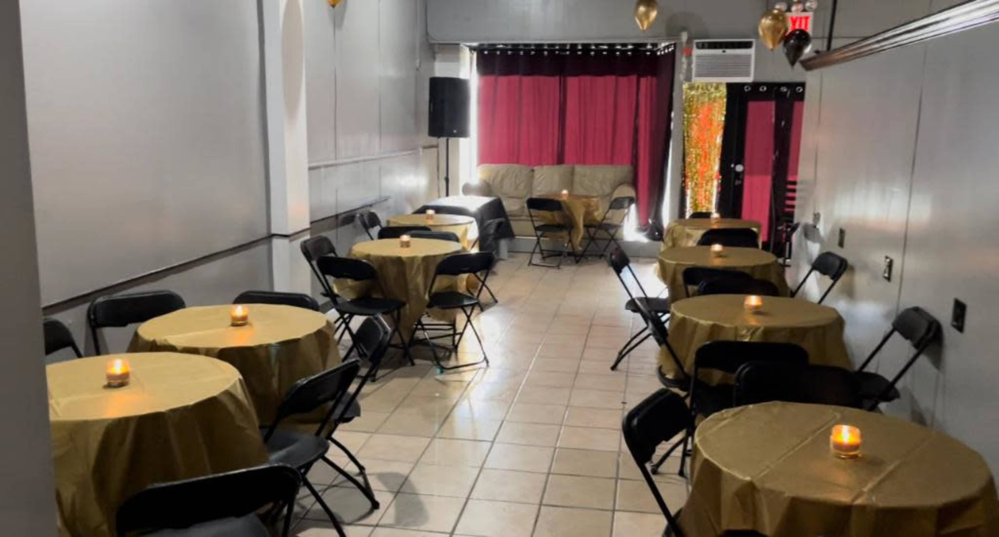 A small event space with gold tablecloths on round tables, each featuring a lit candle, and a backdrop of curtains and an entrance with a decorative element.