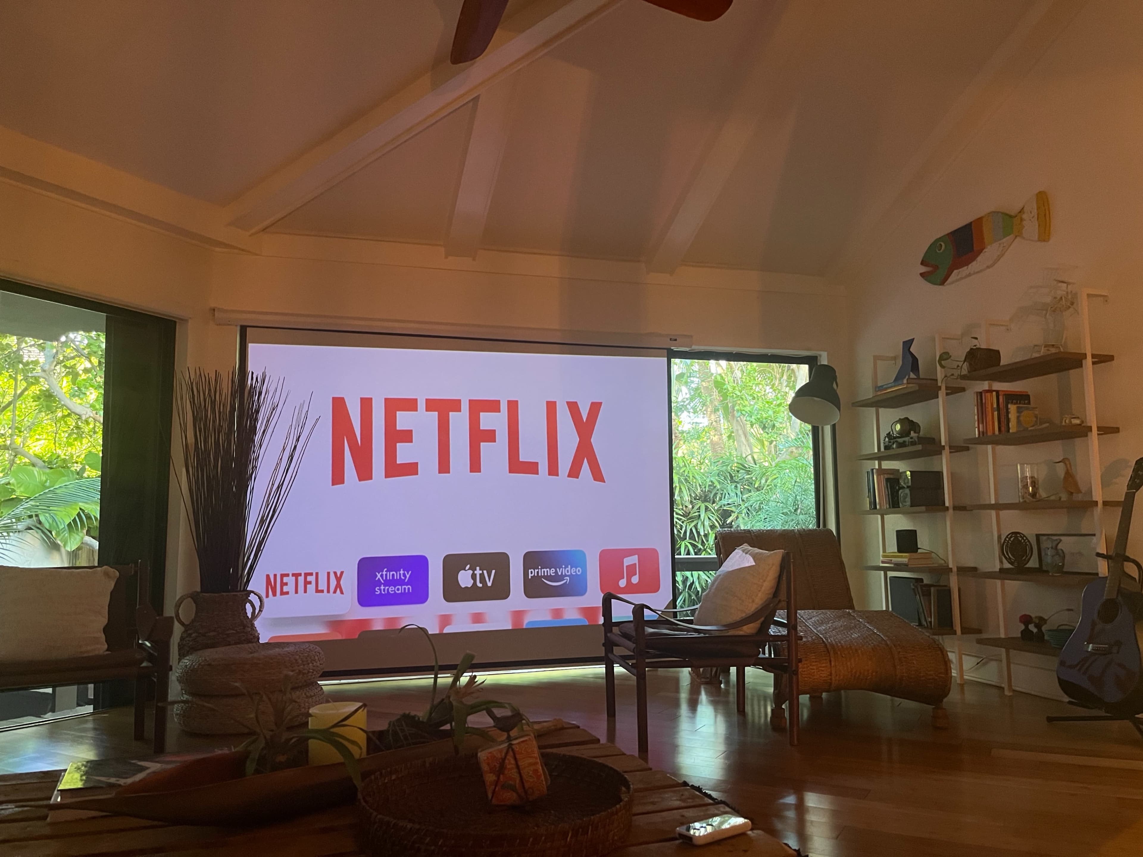 A large screen displaying the Netflix logo is set up in a living room with a tropical view visible through a glass wall.