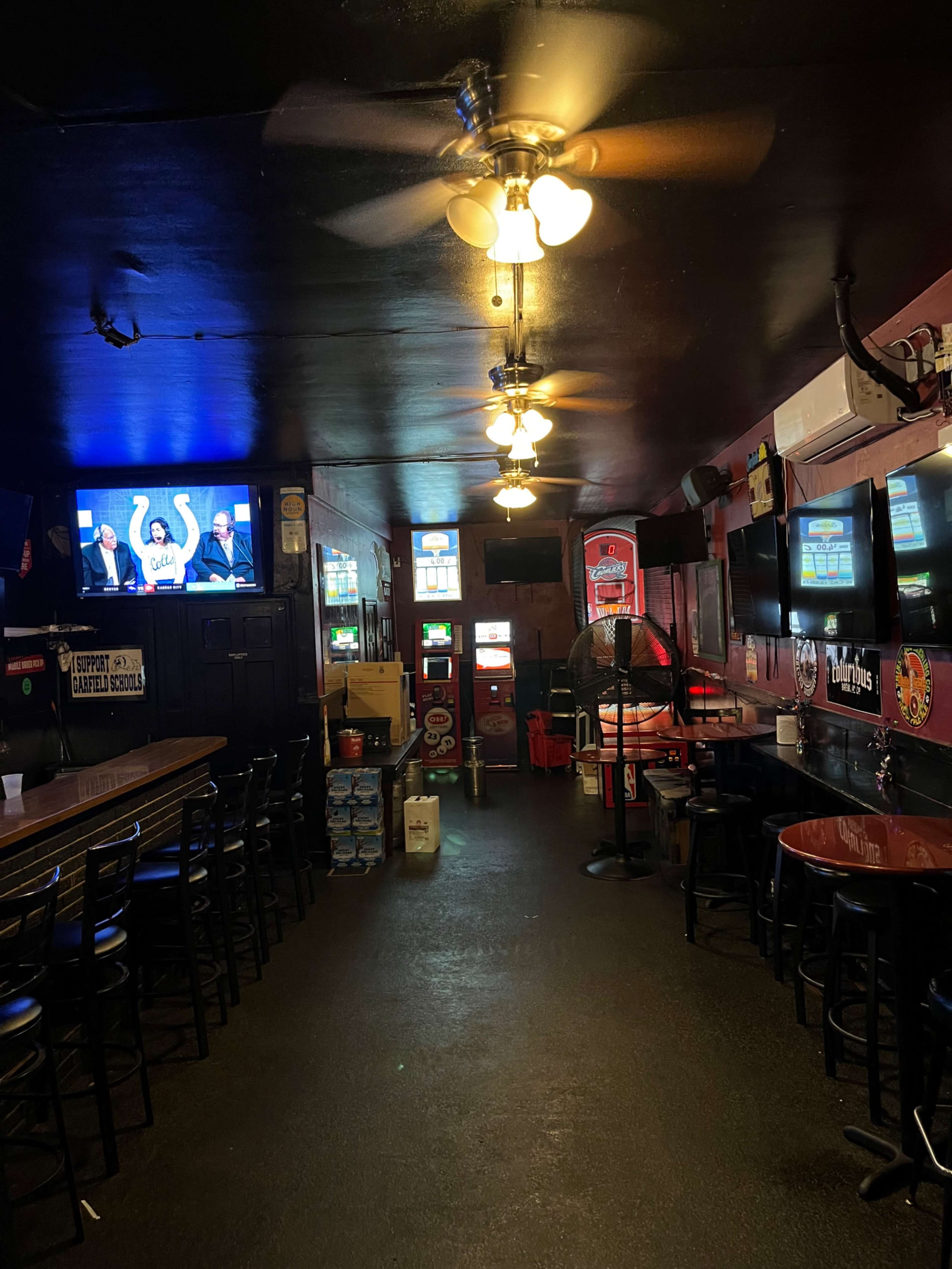The image shows a dimly lit bar interior featuring a row of high-top tables, several video gaming machines, and multiple monitors displaying content along the walls.