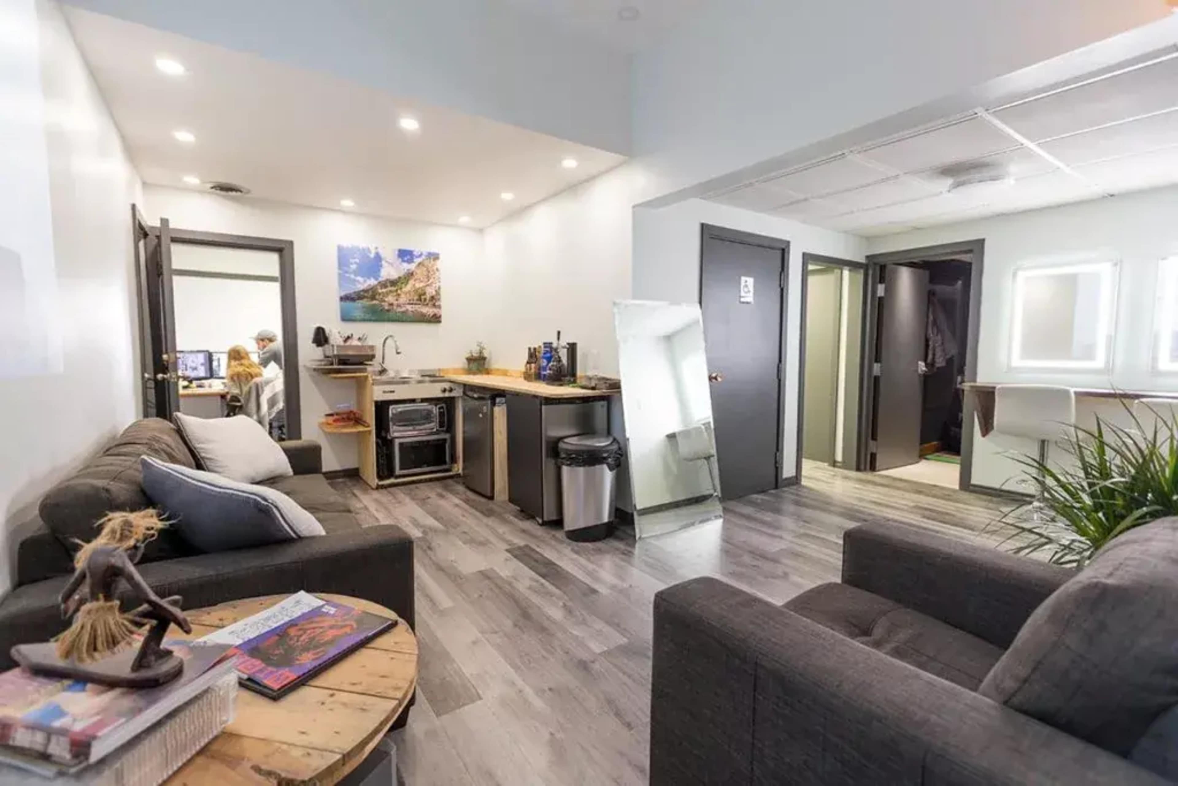 The image shows a modern lounge area with two sofas, a kitchen counter, and an adjacent room with a work station.