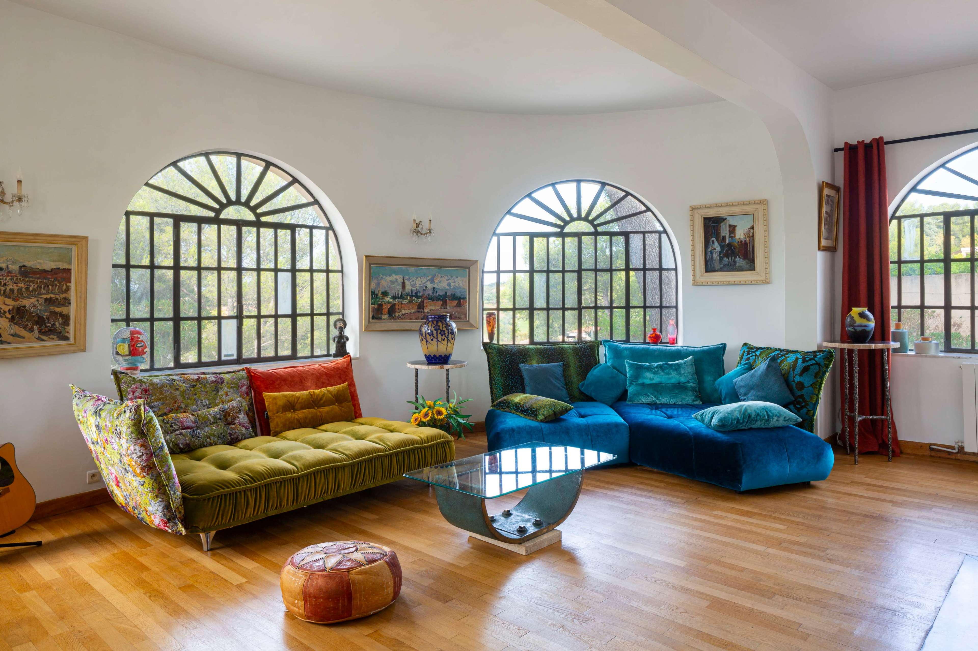 A brightly lit living room features a colorful sofa set, large arched windows, and a wooden floor adorned with decorative pillows and a glass coffee table.