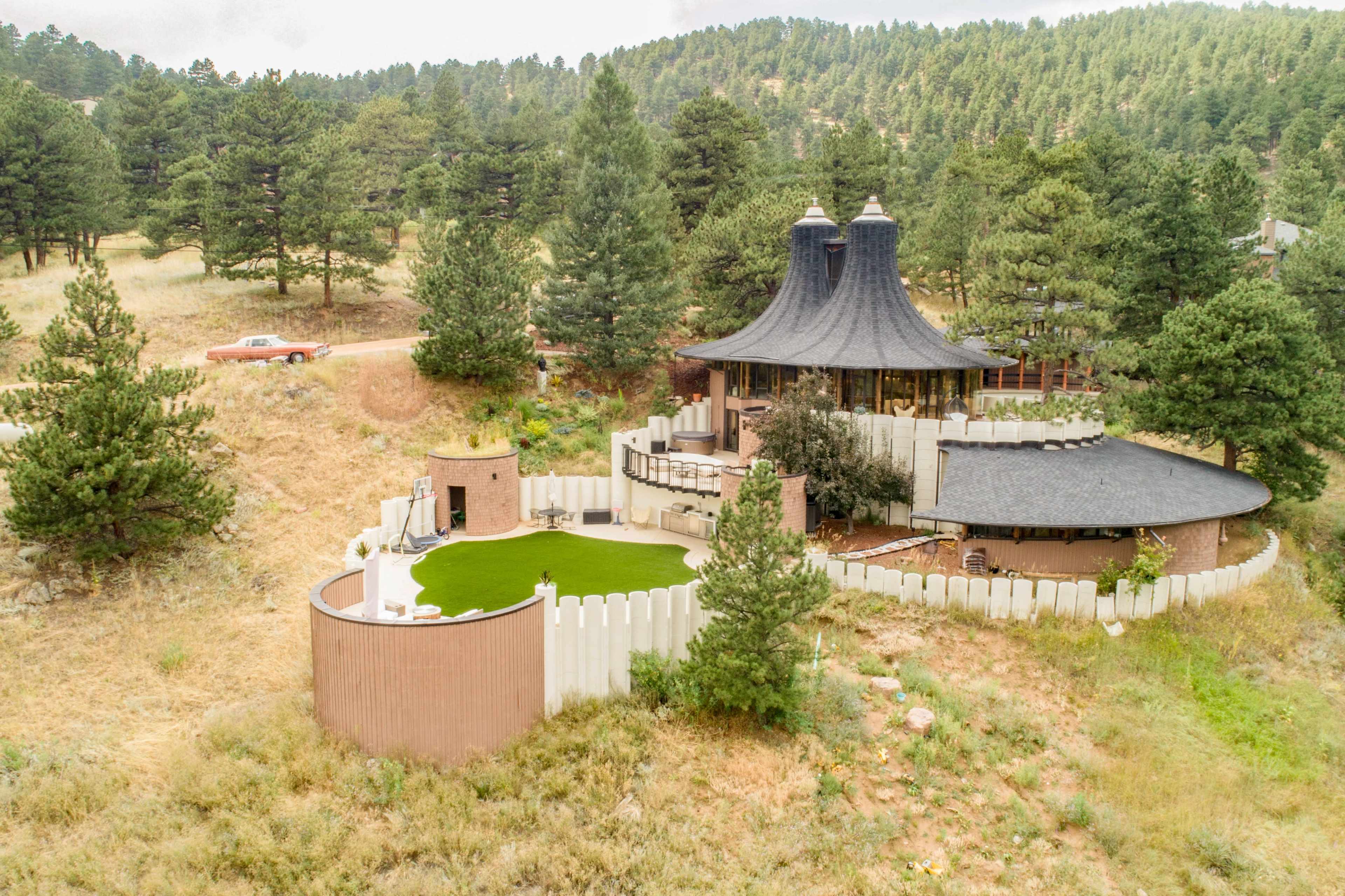 The image shows a unique architectural structure surrounded by trees and grassy terrain, featuring a circular patio and a large lawn area.