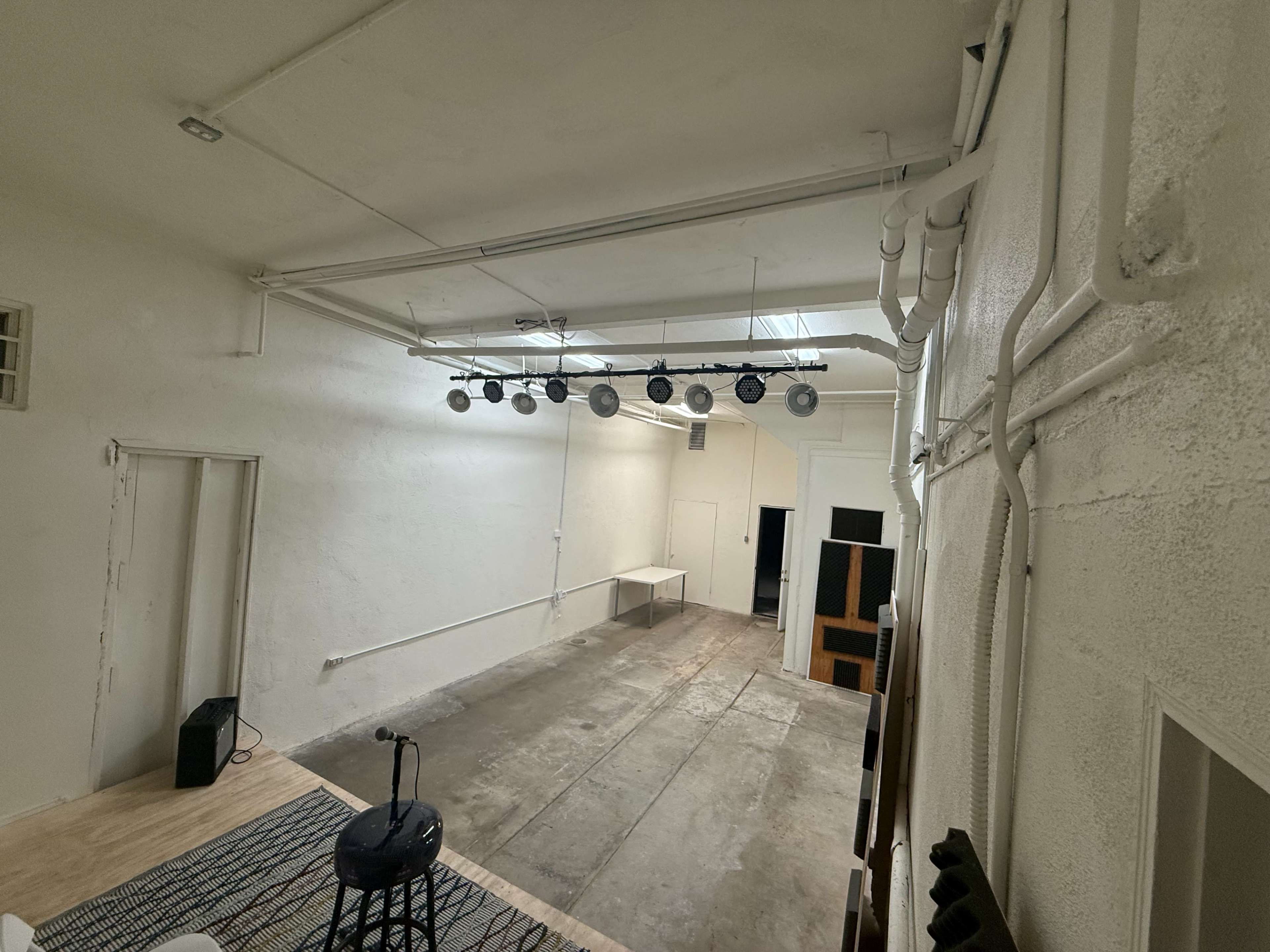 The image shows an empty, well-lit room with overhead lighting fixtures, exposed pipes, and a simple wooden floor.