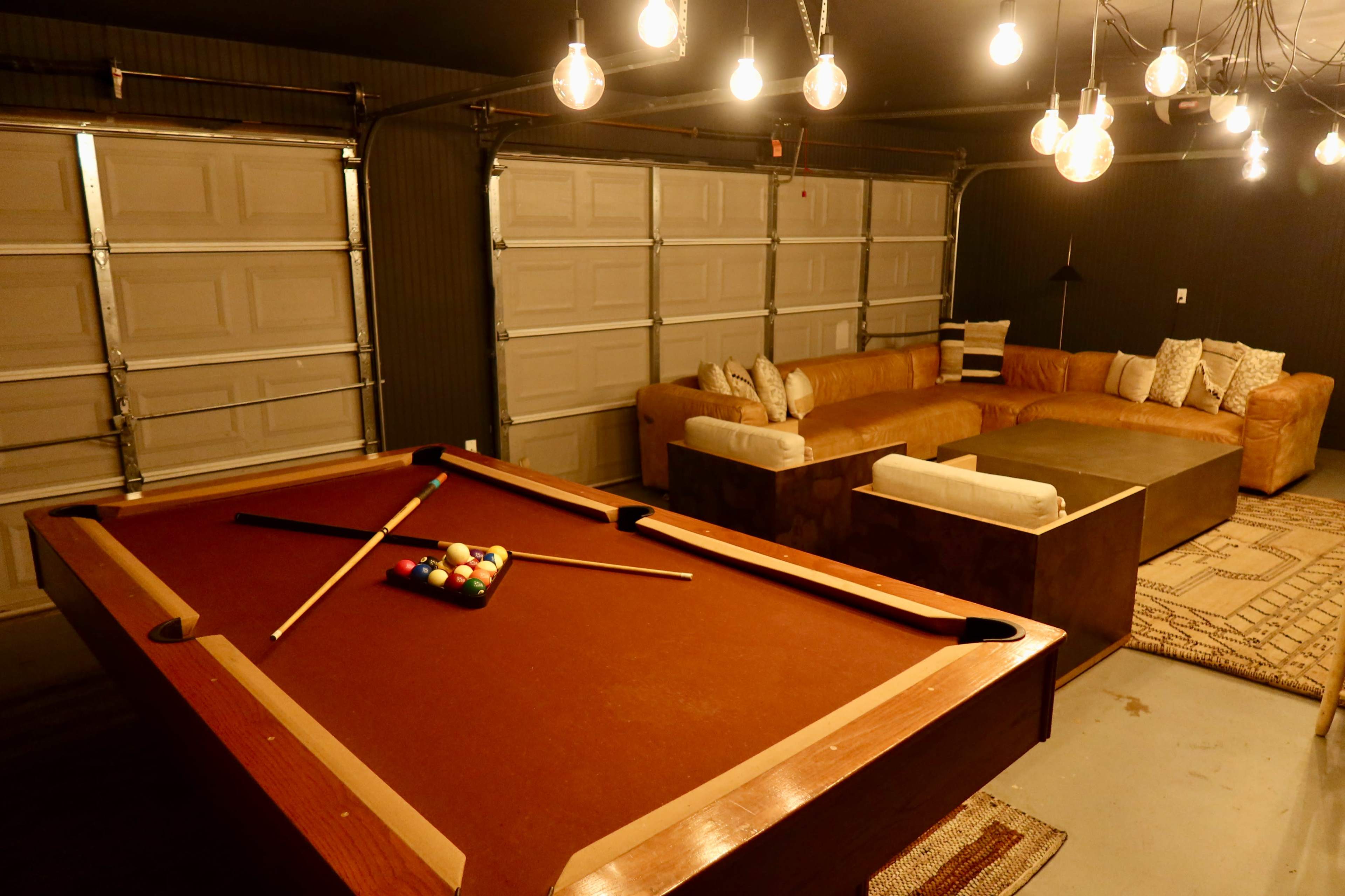 The image shows a game room featuring a pool table with a set of billiard balls arranged on it, alongside a large sectional sofa and warm lighting from hanging bulbs.