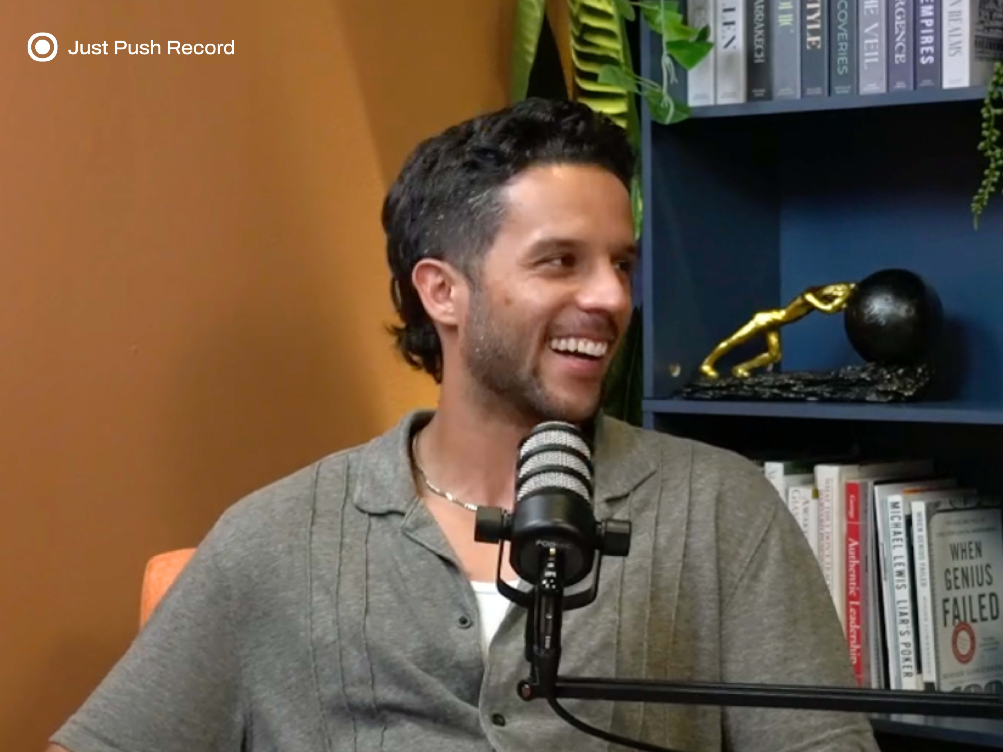 A man sitting in a chair smiles while speaking into a microphone in front of a colorful bookshelf.