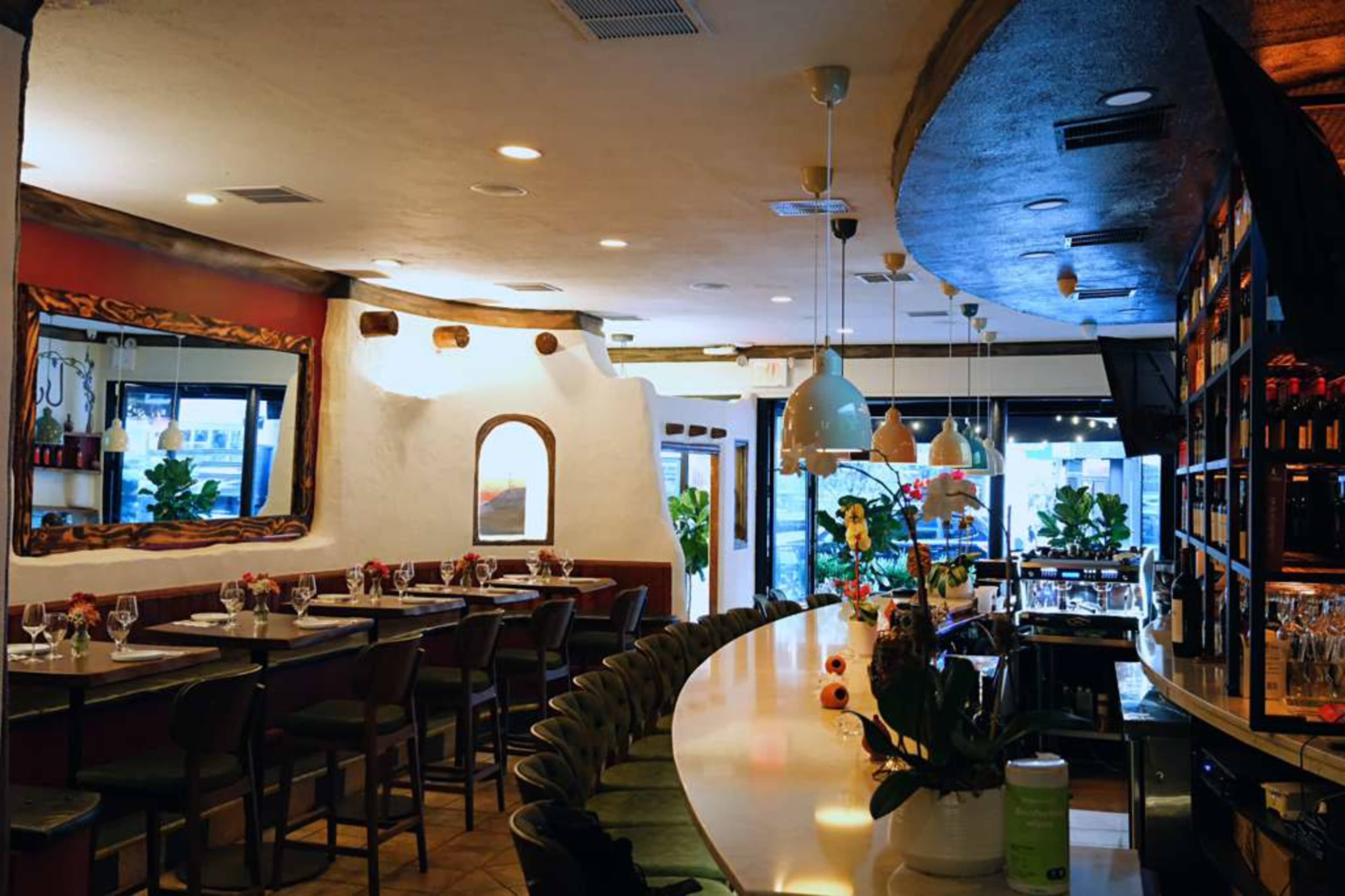 The interior of a restaurant features a curved bar with high stools, elegant lighting, and plants, surrounded by walls decorated with mirrors and art.