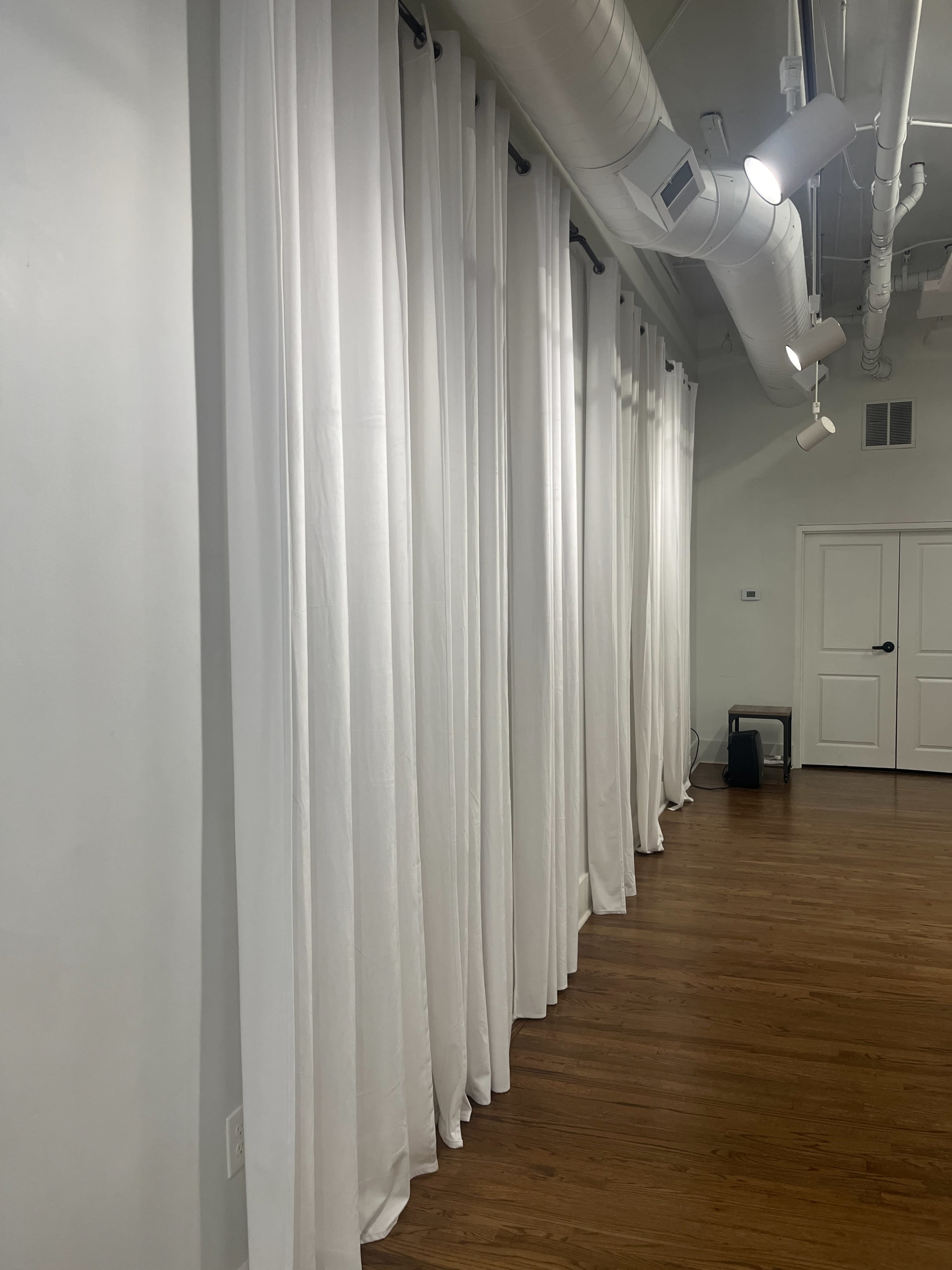 The image shows a series of white curtains hanging along a wall in a room with wooden flooring and ceiling ducts.