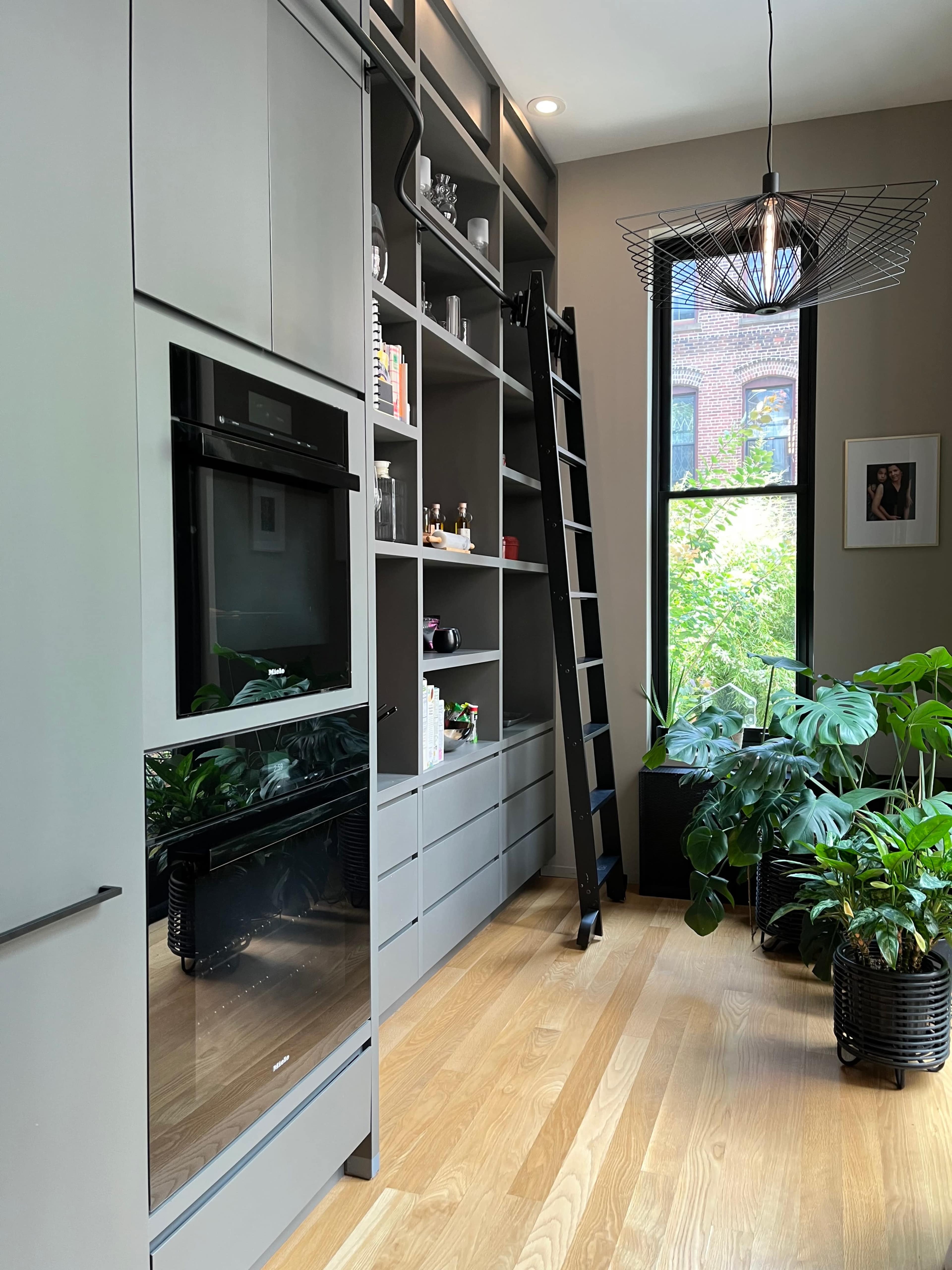 A modern kitchen featuring gray cabinetry, an oven, a tall shelving unit, a black ladder, and large windows with green plants visible outside.