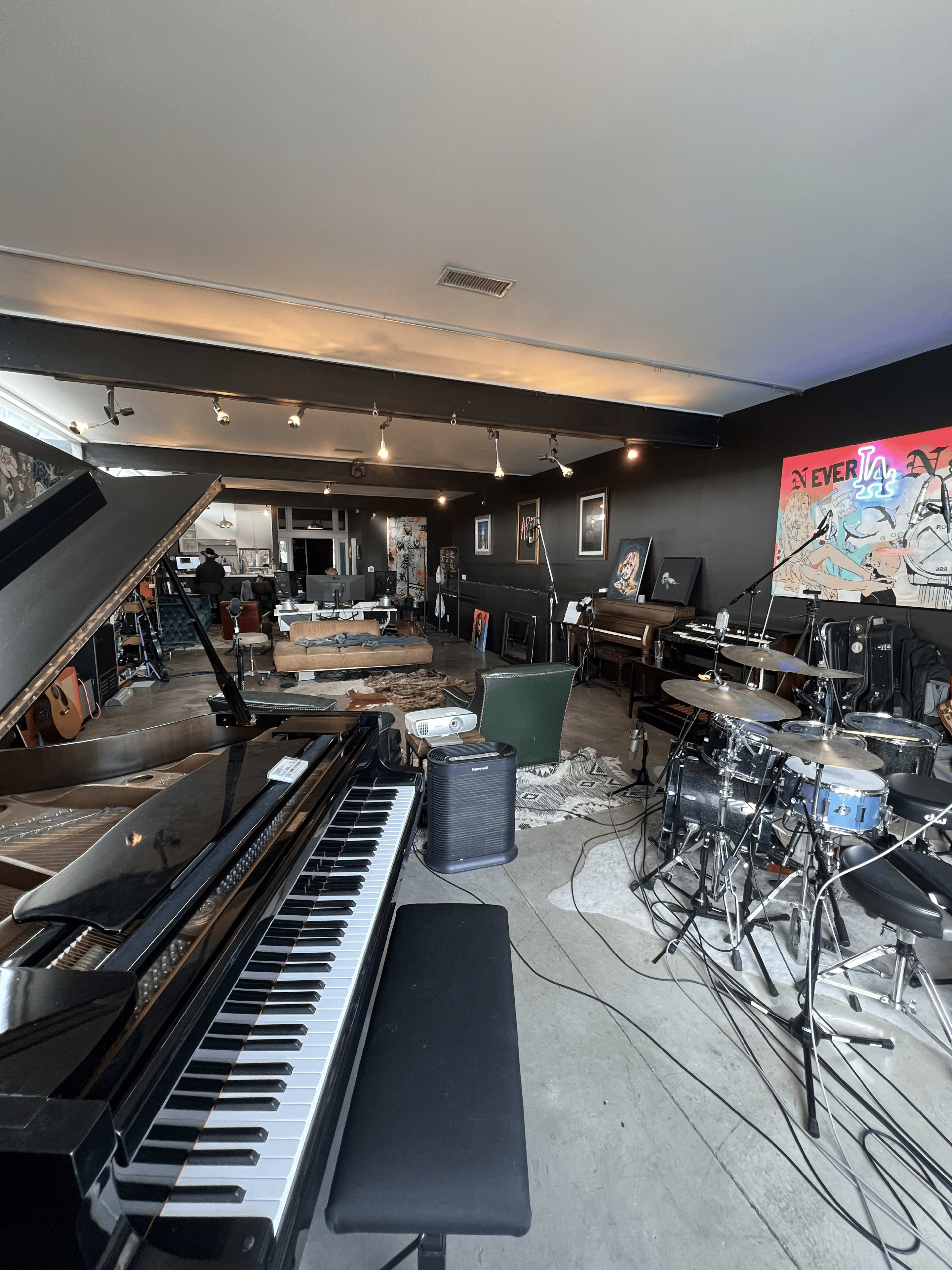 The image shows a spacious music studio featuring a grand piano, a drum set, various instruments, and a dark-painted wall adorned with artwork.