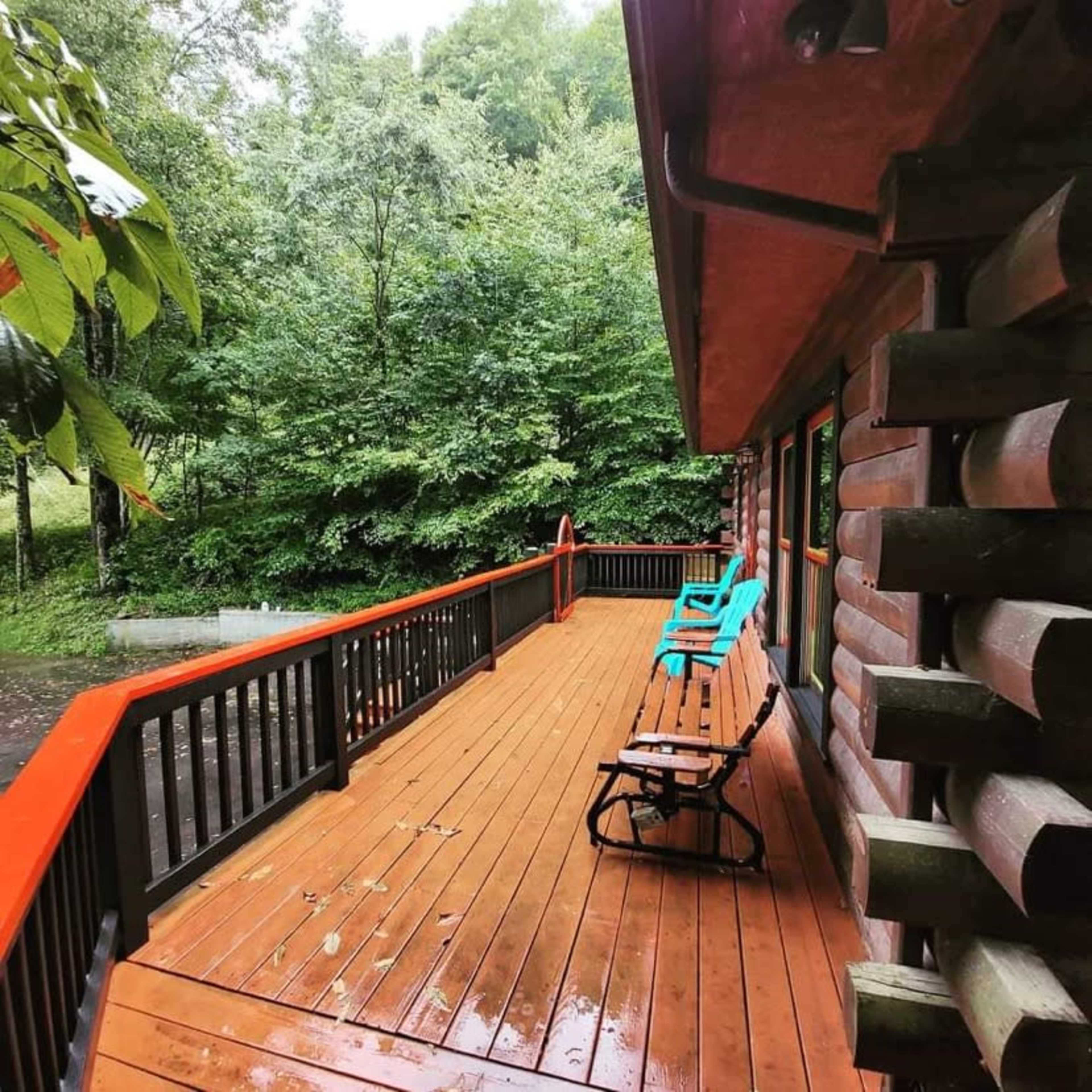 A wooden deck with orange railings and two turquoise chairs overlooks a lush, green wooded area.