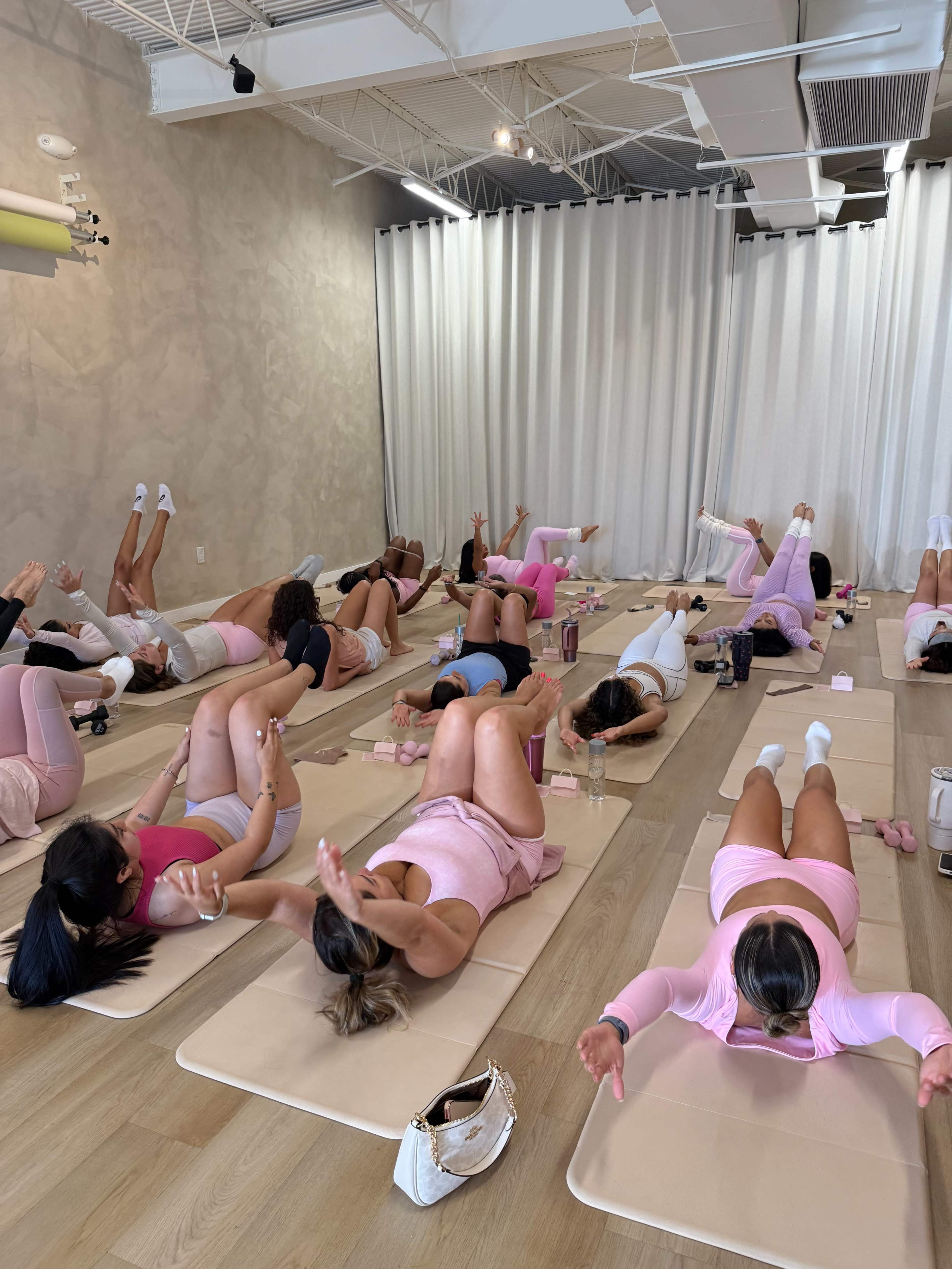 A group of people in athletic attire is participating in a fitness class on mats in a spacious studio with light-colored walls and floor.