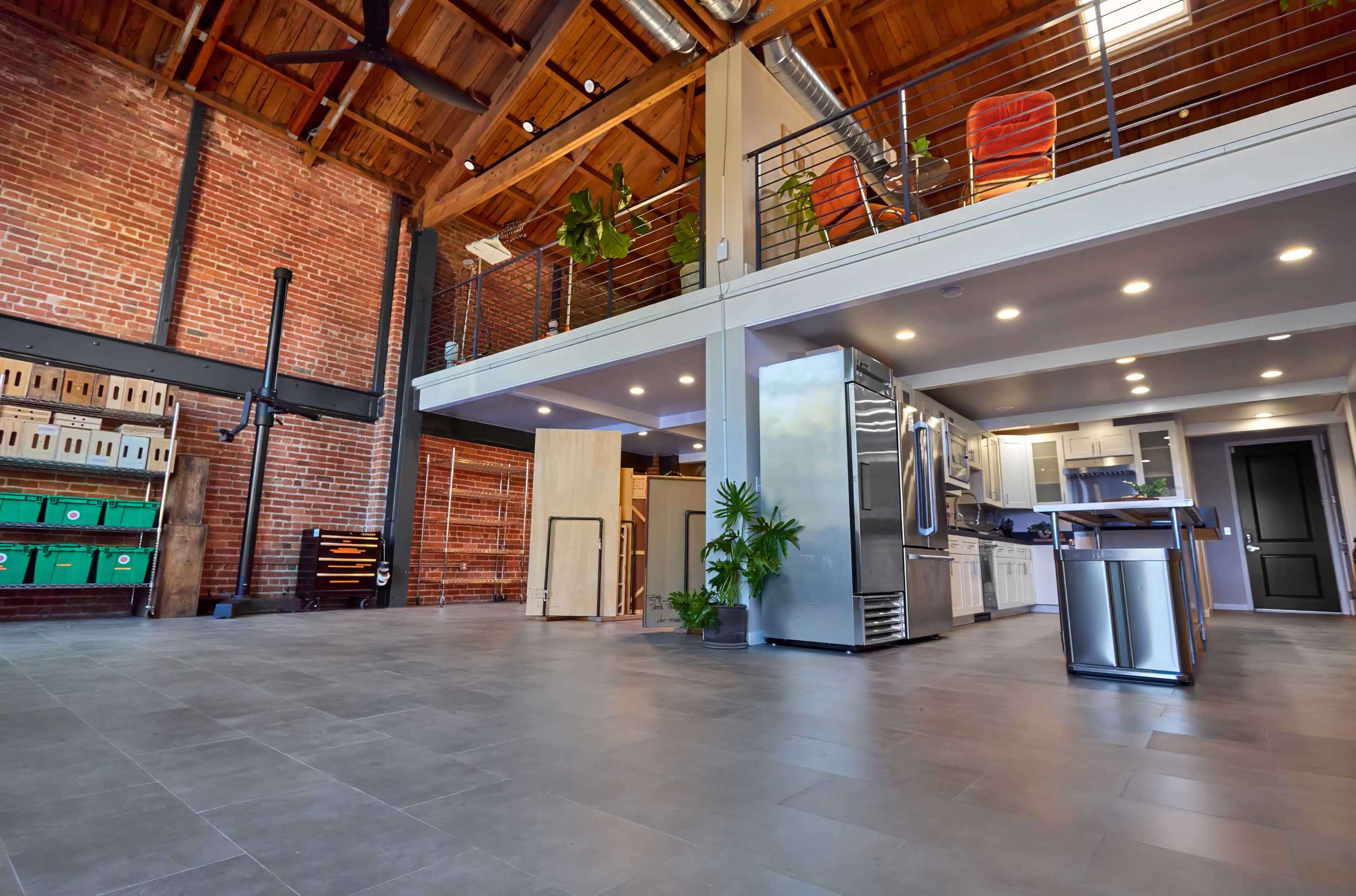 The image shows a spacious, open-concept industrial-style interior featuring brick walls, high ceilings with wooden beams, and a modern kitchen area.