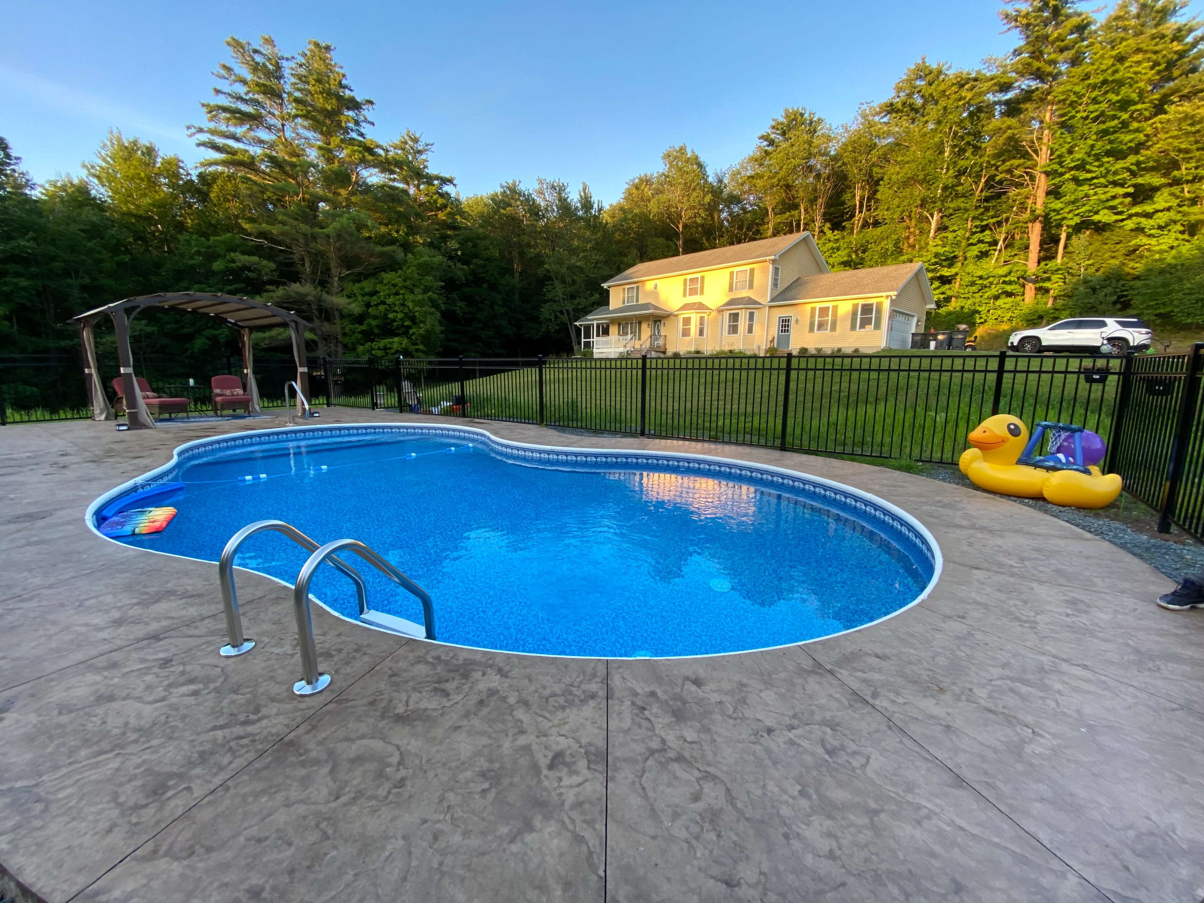 A blue swimming pool with ladders, surrounded by a concrete patio, features a yellow inflatable duck and a house in the background, all set against a wooded area.