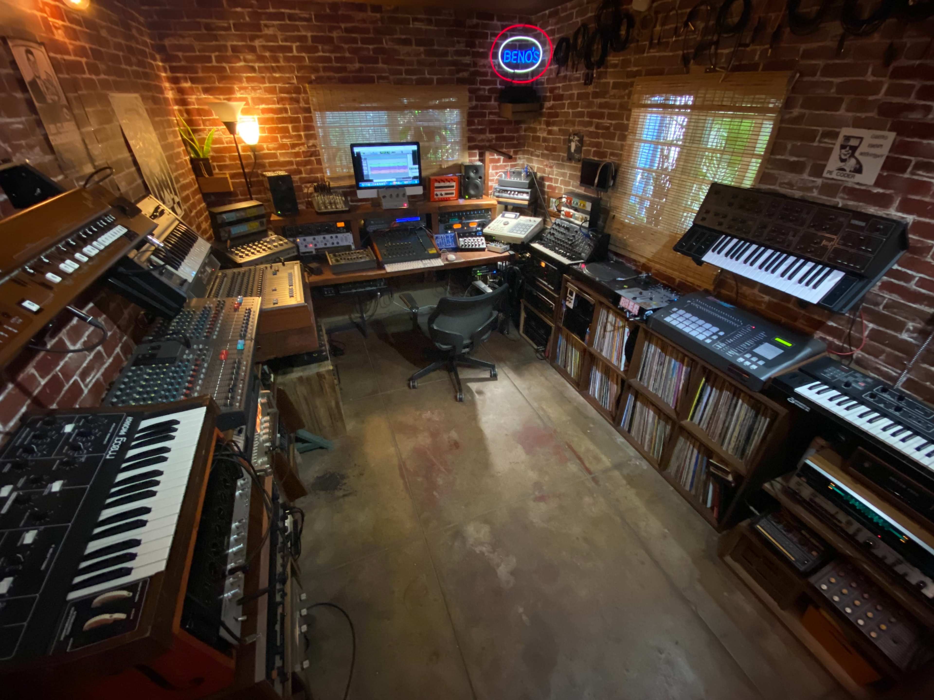 A recording studio filled with various synthesizers, keyboards, and equipment, arranged around a central workspace with wooden shelves and a neon sign.
