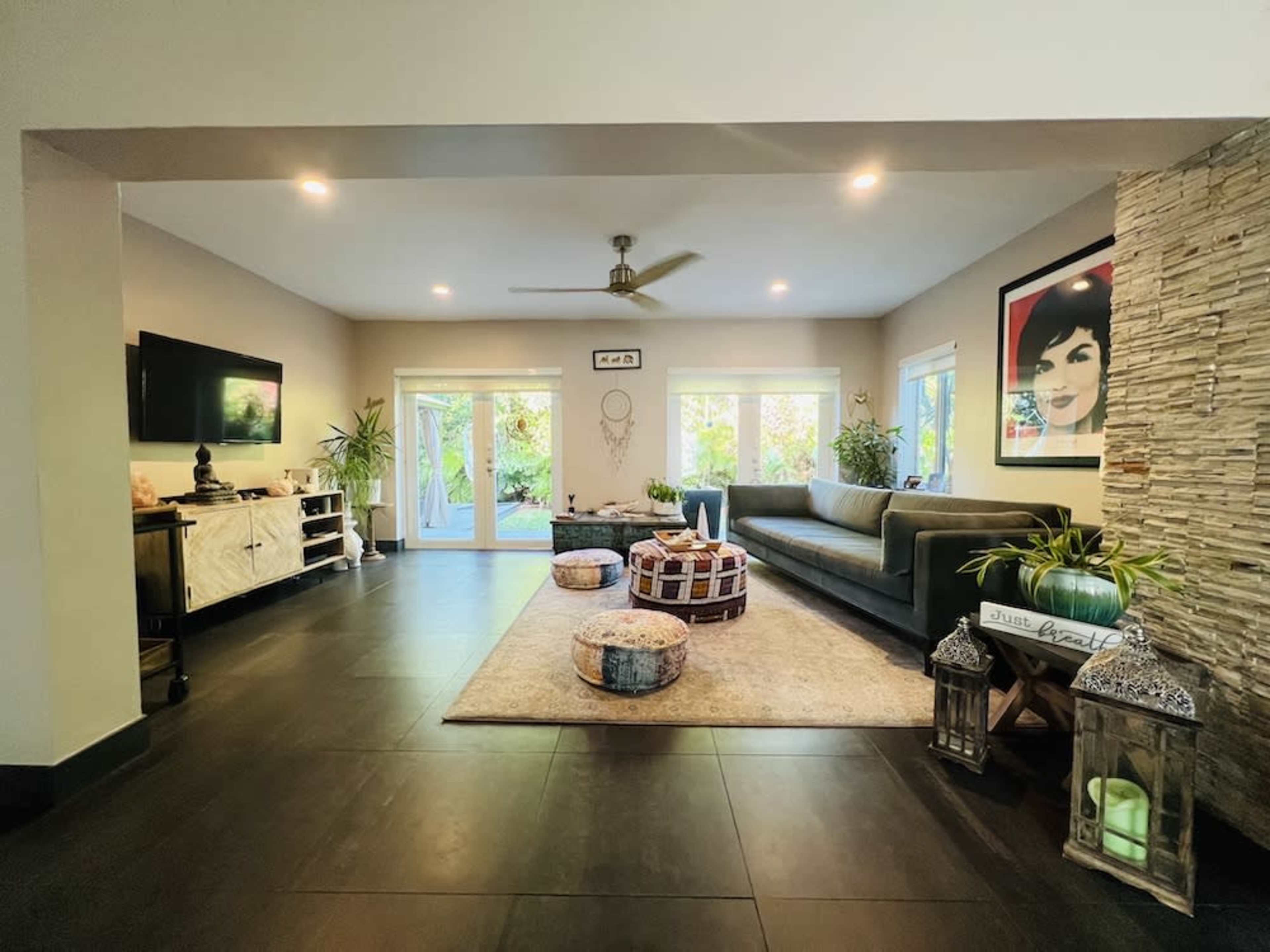 A modern living room with a dark tiled floor, a gray sofa, decorative poufs, and large windows showcasing greenery outside.
