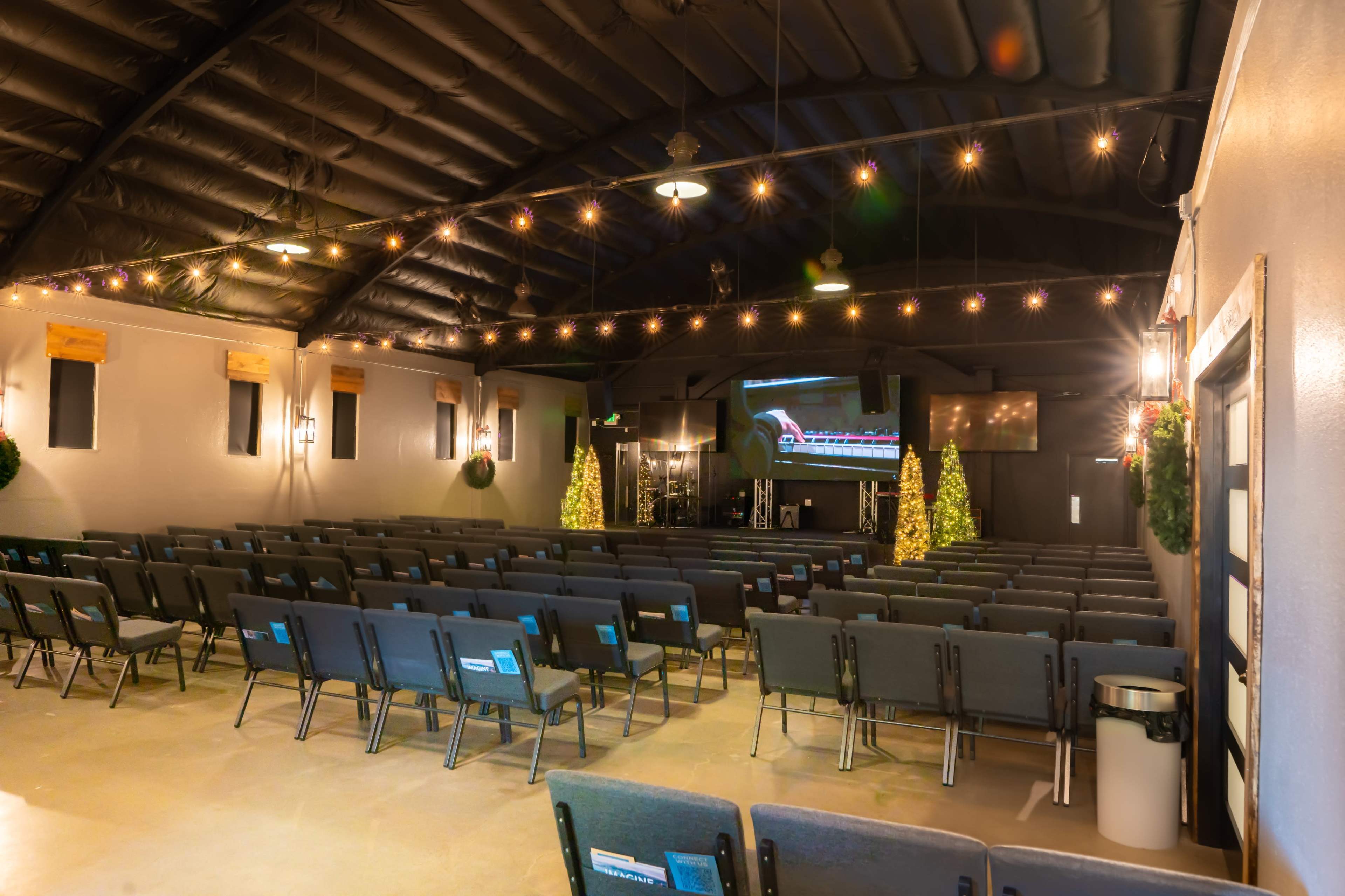 An empty indoor auditorium with rows of chairs, a large screen at the front, and decorative lights and greenery.