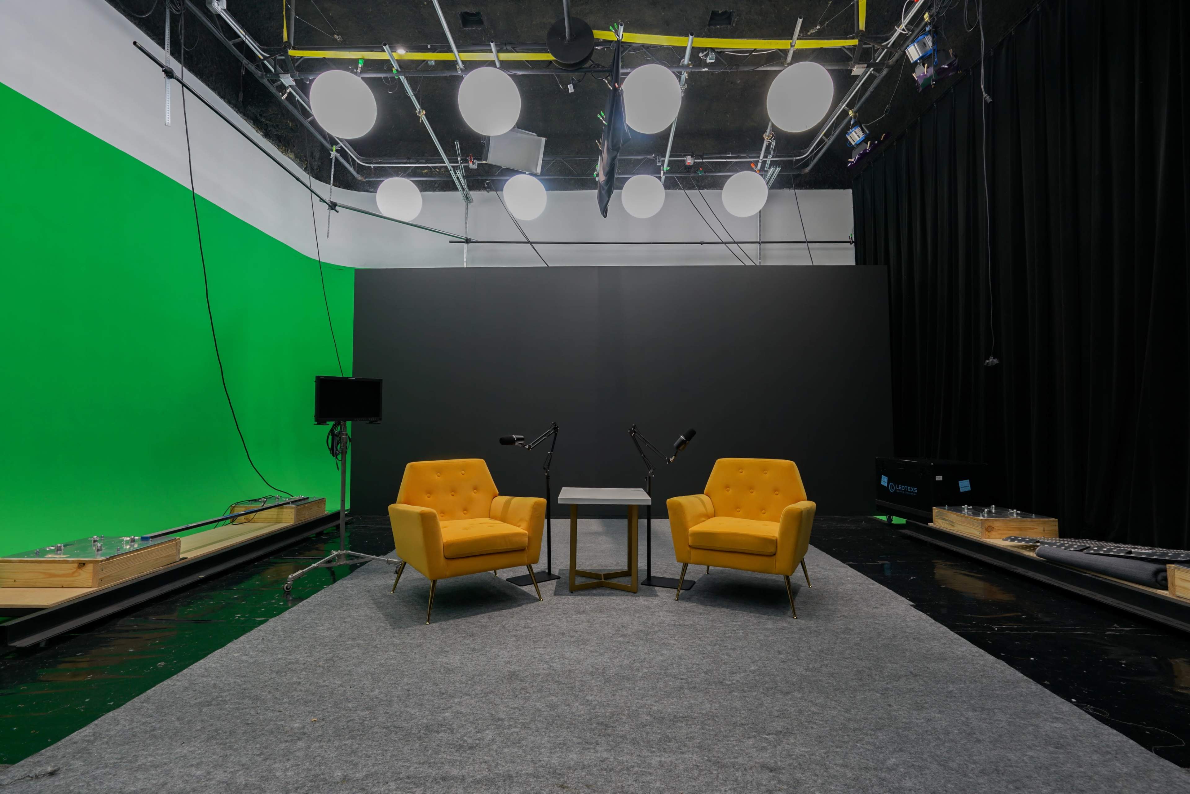 The image shows a video production studio featuring two yellow chairs, a small table between them, and green and black backdrop walls.