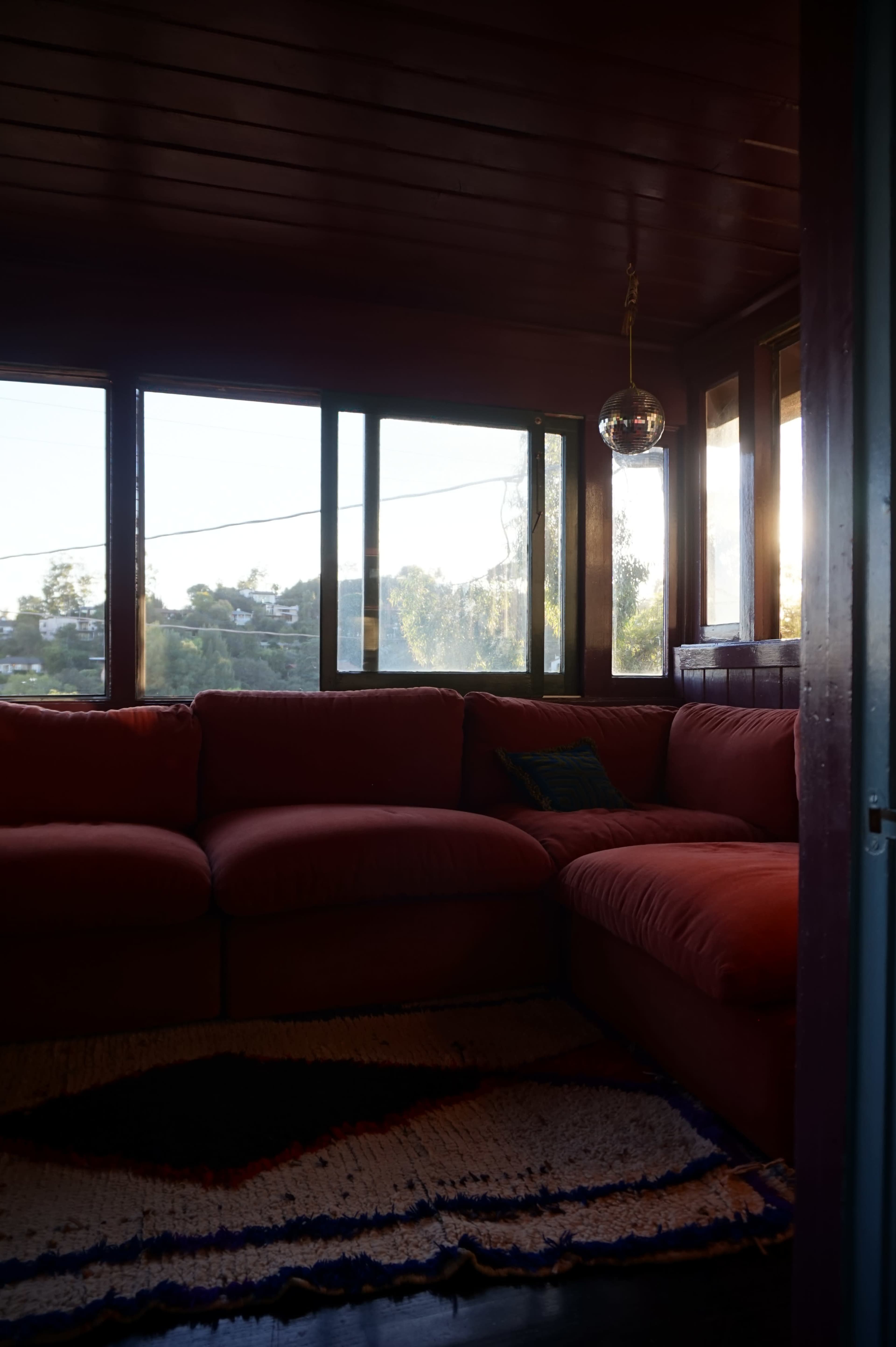 The image shows a corner of a cozy room featuring a red sectional sofa, large windows with a view of greenery outside, and a hanging light fixture.