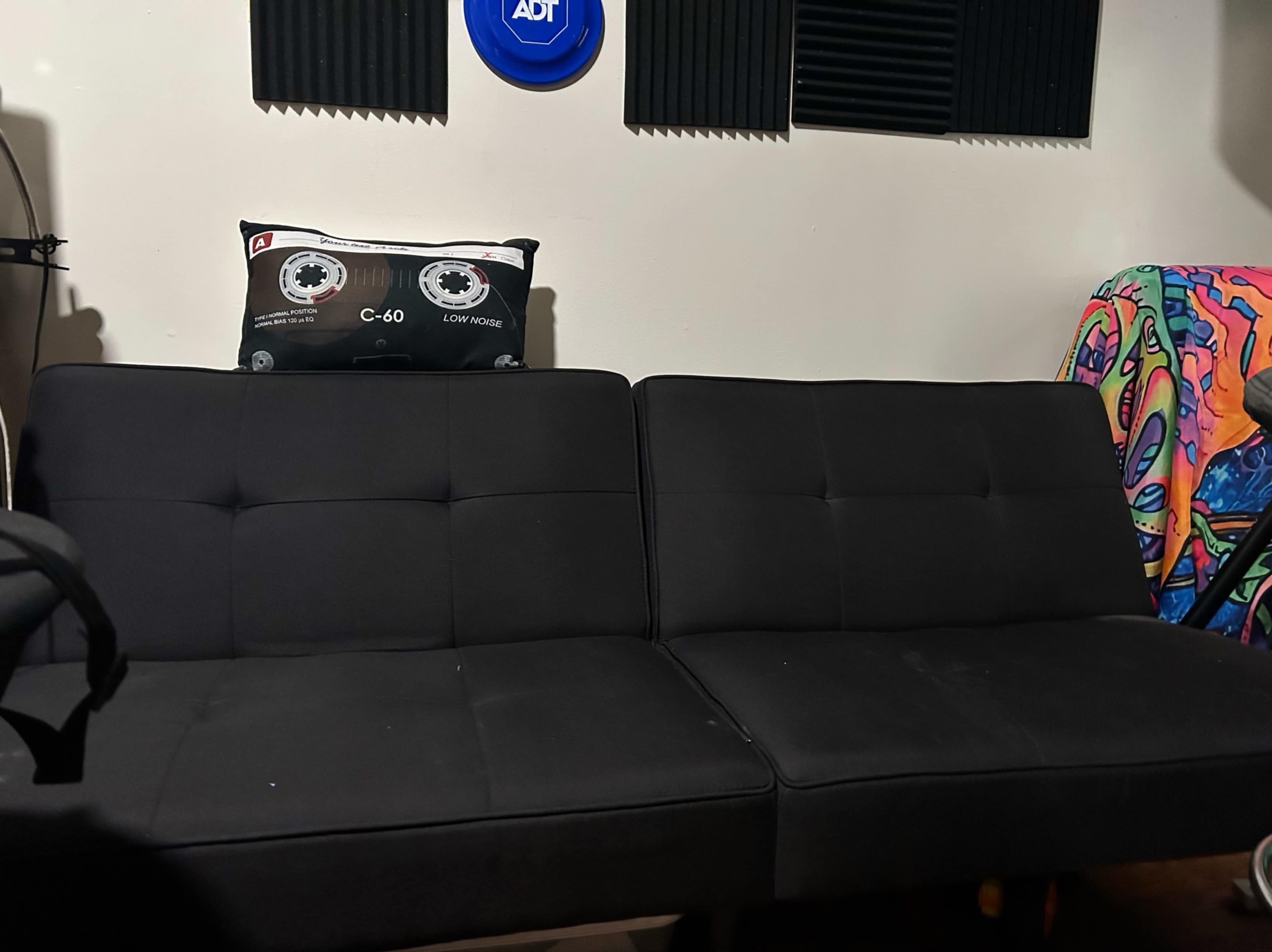 The image shows a black sofa with a decorative pillow featuring a cassette tape design, positioned against a wall with sound-absorbing panels.