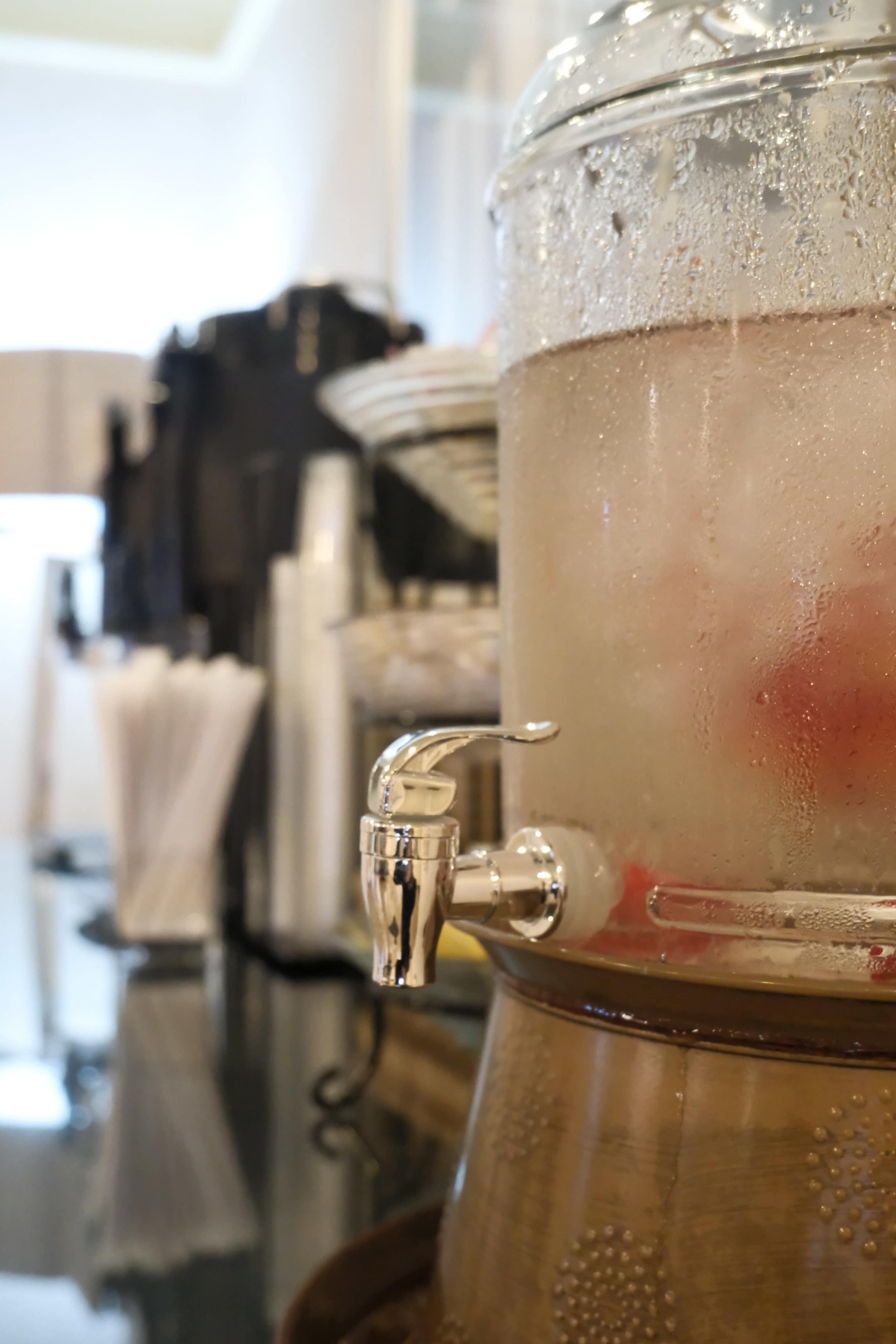 A glass beverage dispenser is shown filled with a clear liquid and sitting on a wooden base, with a metallic spout for pouring.