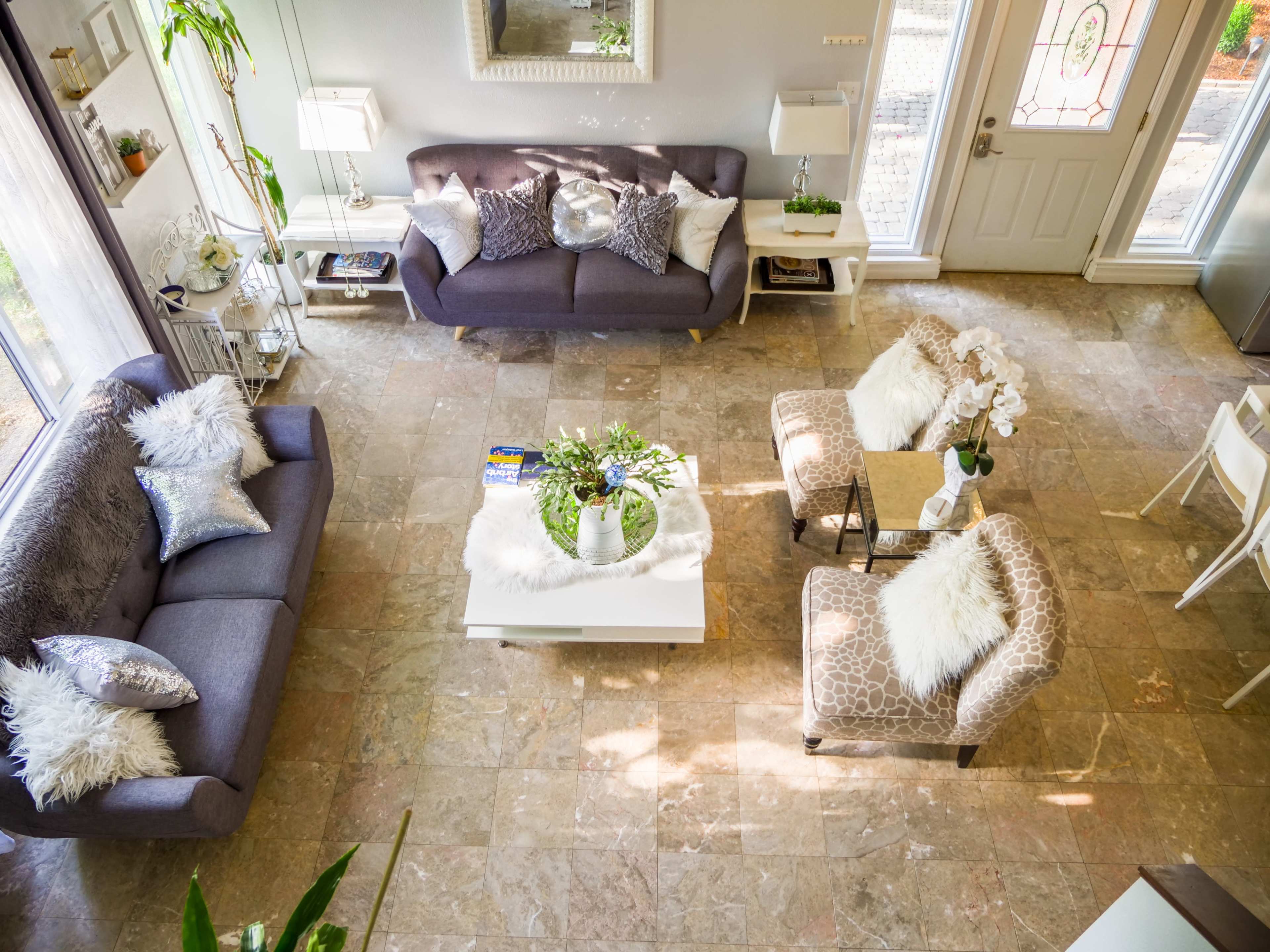 A well-lit living room with two sofas, a coffee table, and decorative plants arranged on a tiled floor.