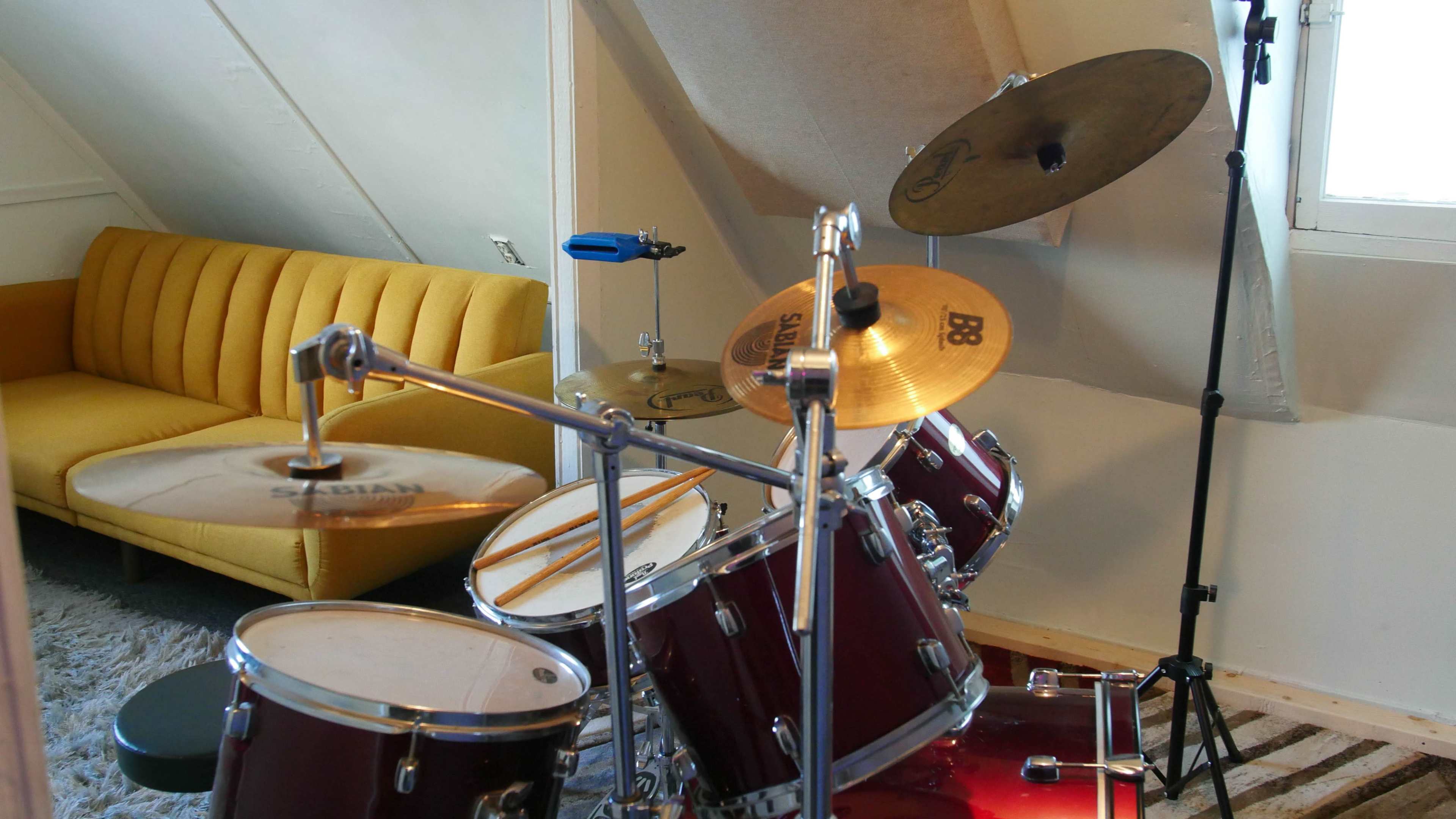 A red drum set is positioned in an attic room beside a yellow couch and a window.