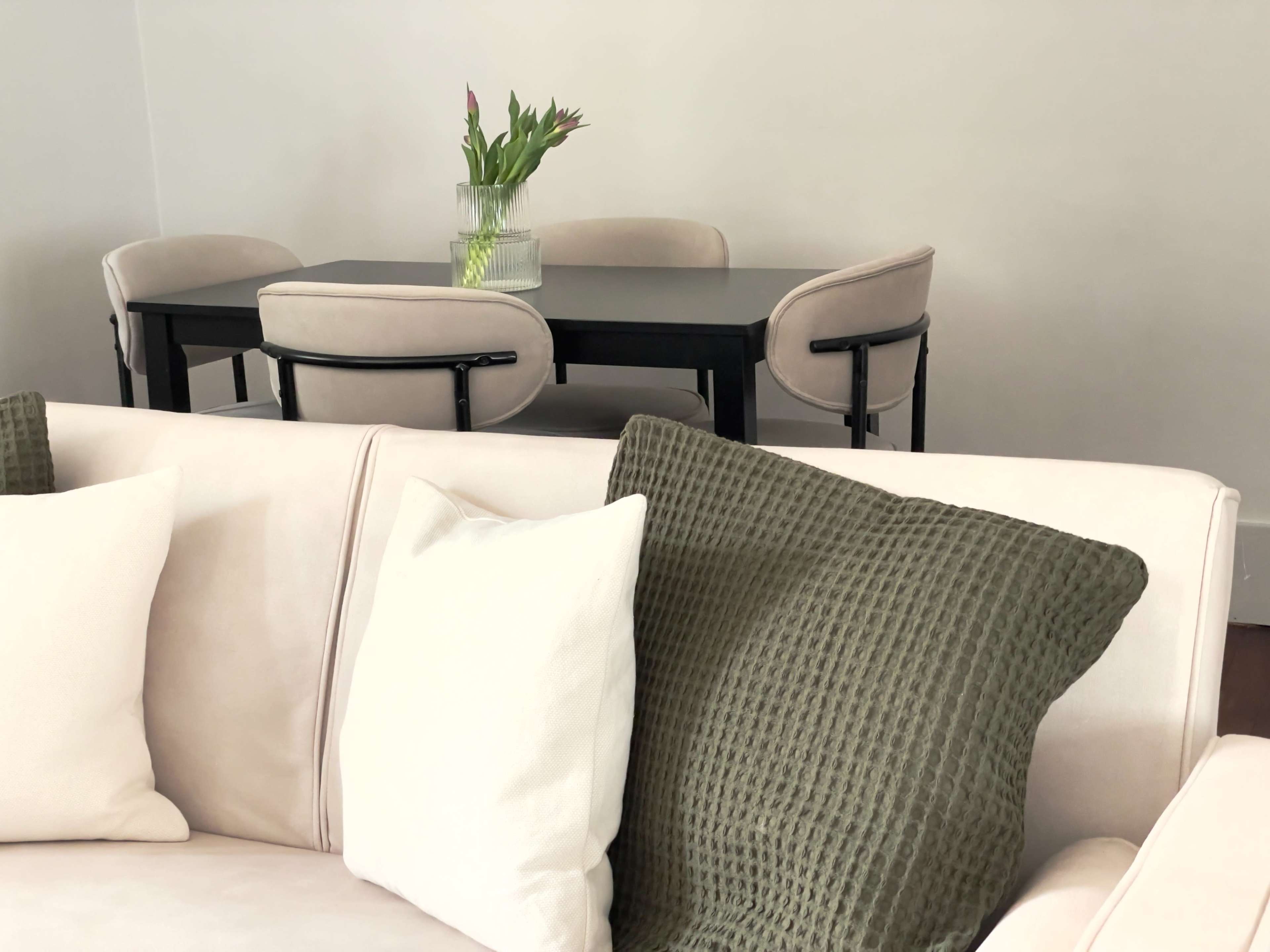 A light-colored couch with multiple cushions is positioned in front of a black dining table and four chairs, complemented by a vase of flowers on the table.