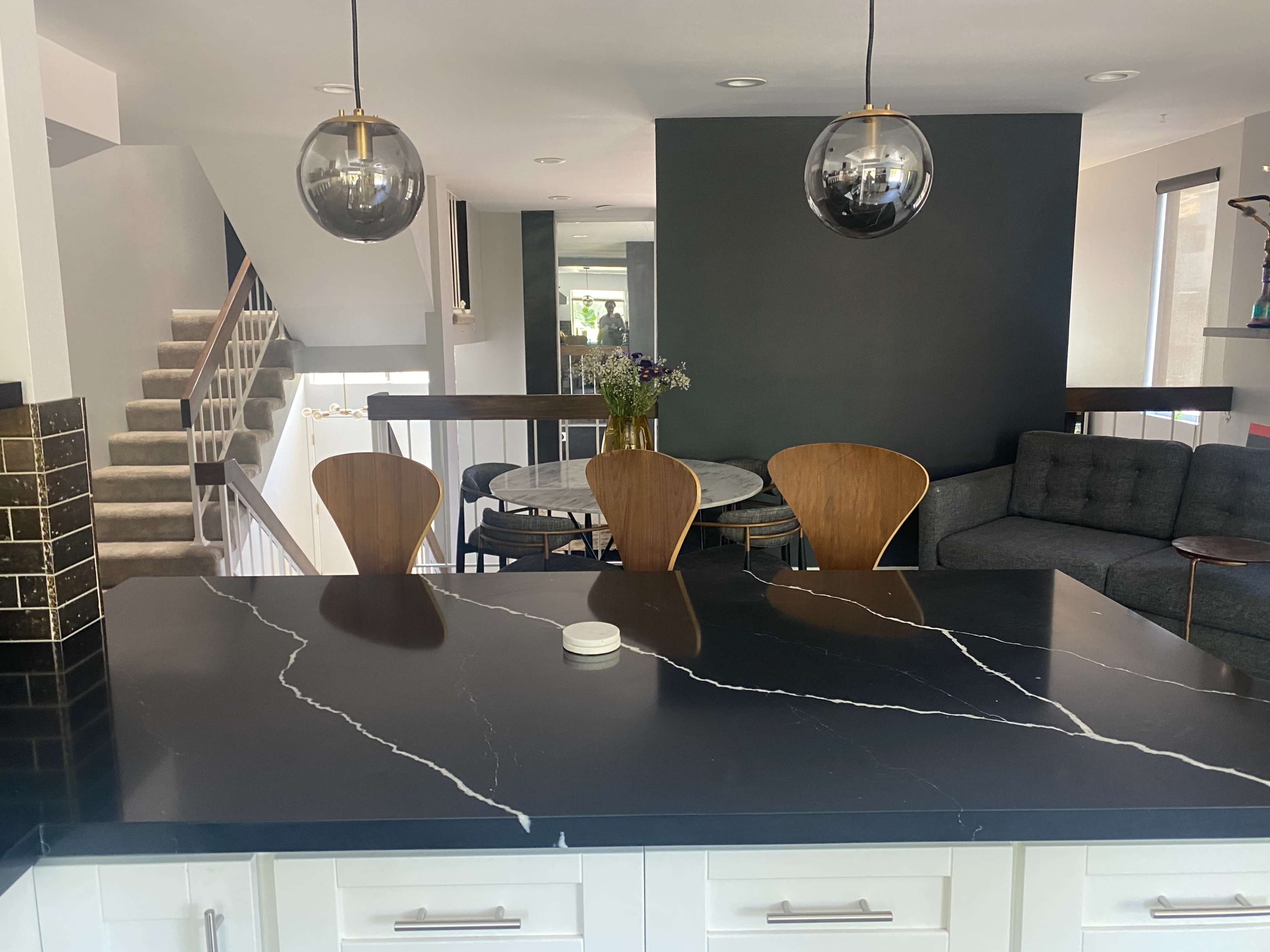 A modern kitchen features a black marble countertop with three wooden chairs and a dining table in the background, illuminated by round pendant lights.