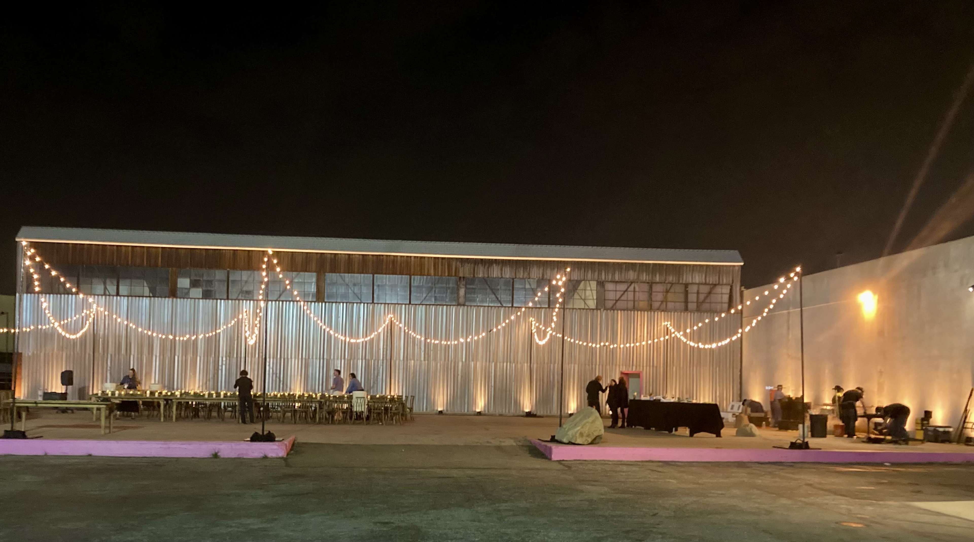 A large industrial space is decorated with string lights and set up for an event, featuring several tables arranged in a banquet style.