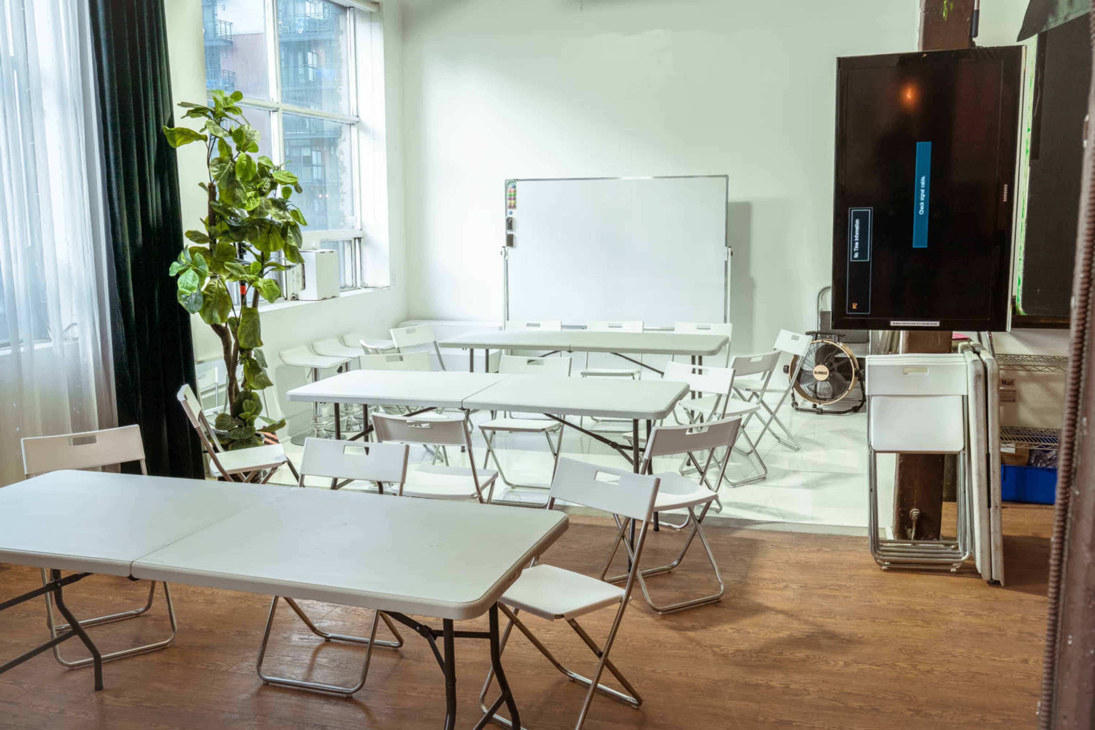 A bright classroom is arranged with several folding tables and chairs, alongside a whiteboard and a television on the wall.