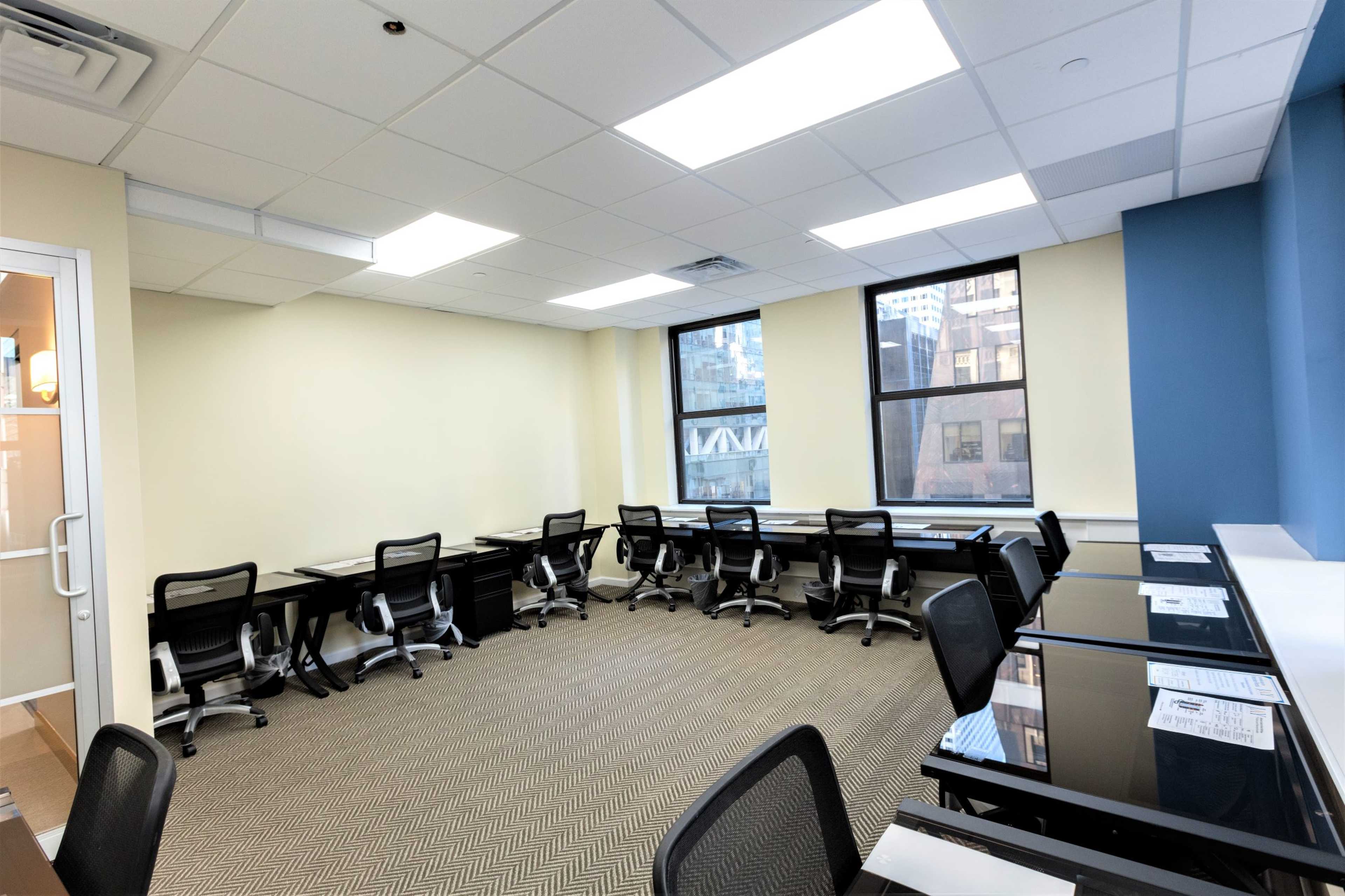 The image depicts a modern office space with several desks and office chairs arranged along two walls, featuring large windows that allow natural light to enter.