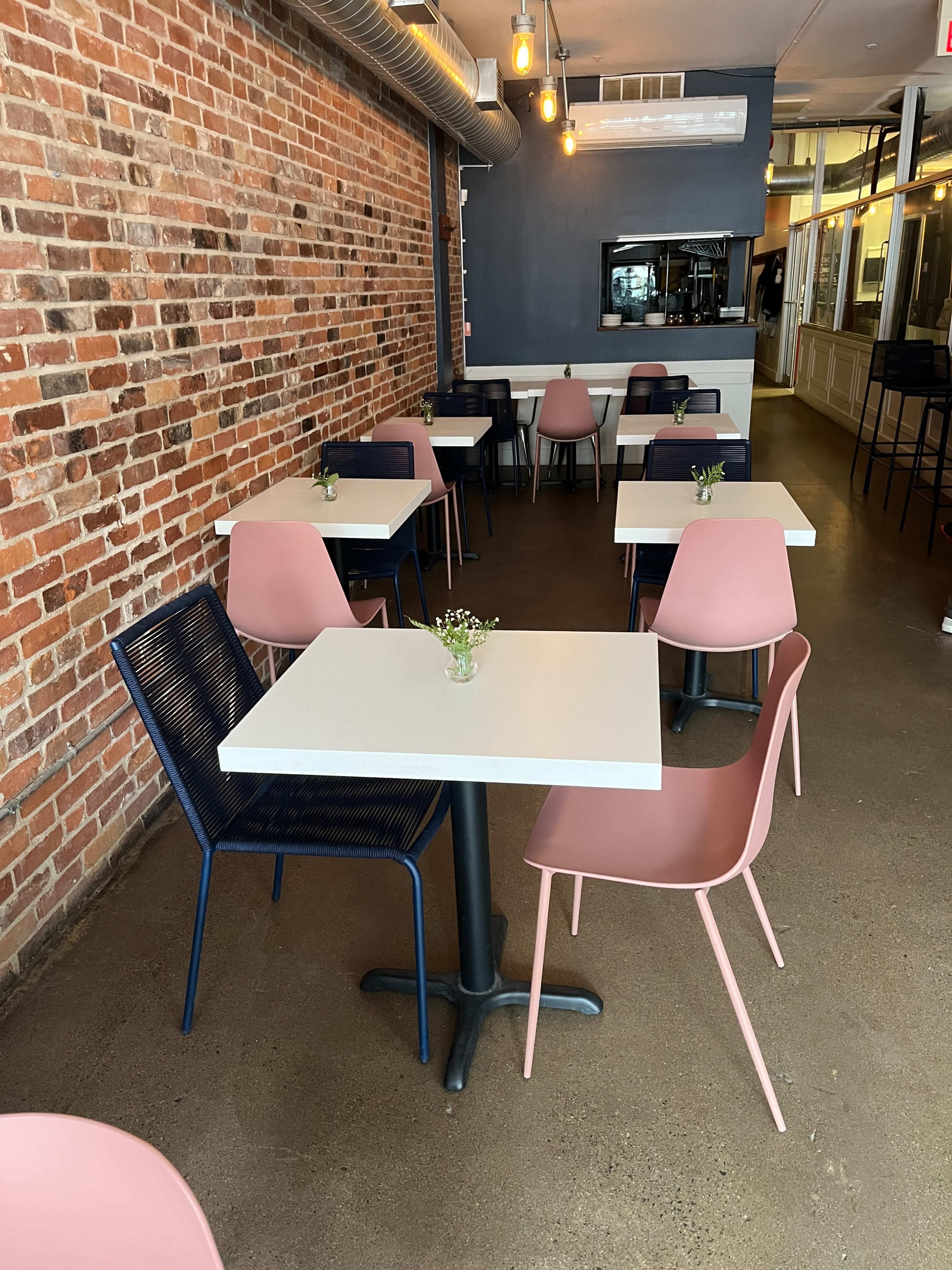 A modern restaurant interior featuring several tables with white tops and pink and navy chairs, along a brick wall with pendant lights above.