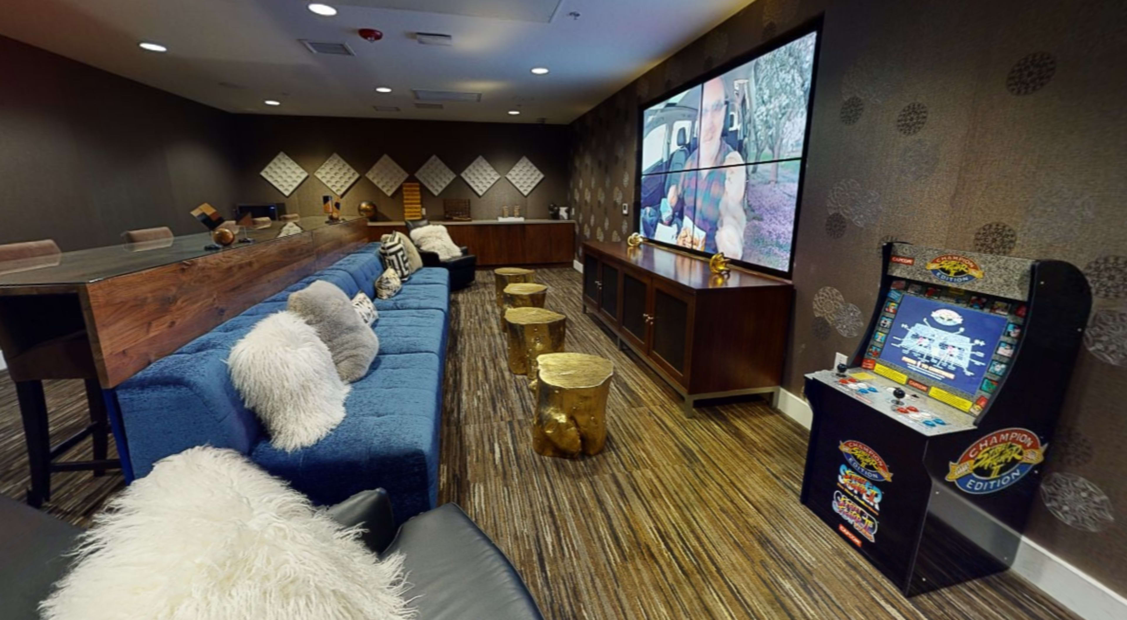 A stylish lounge area with a blue sofa, wooden bar, gold stools, and an arcade game in front of a large screen displaying a video.