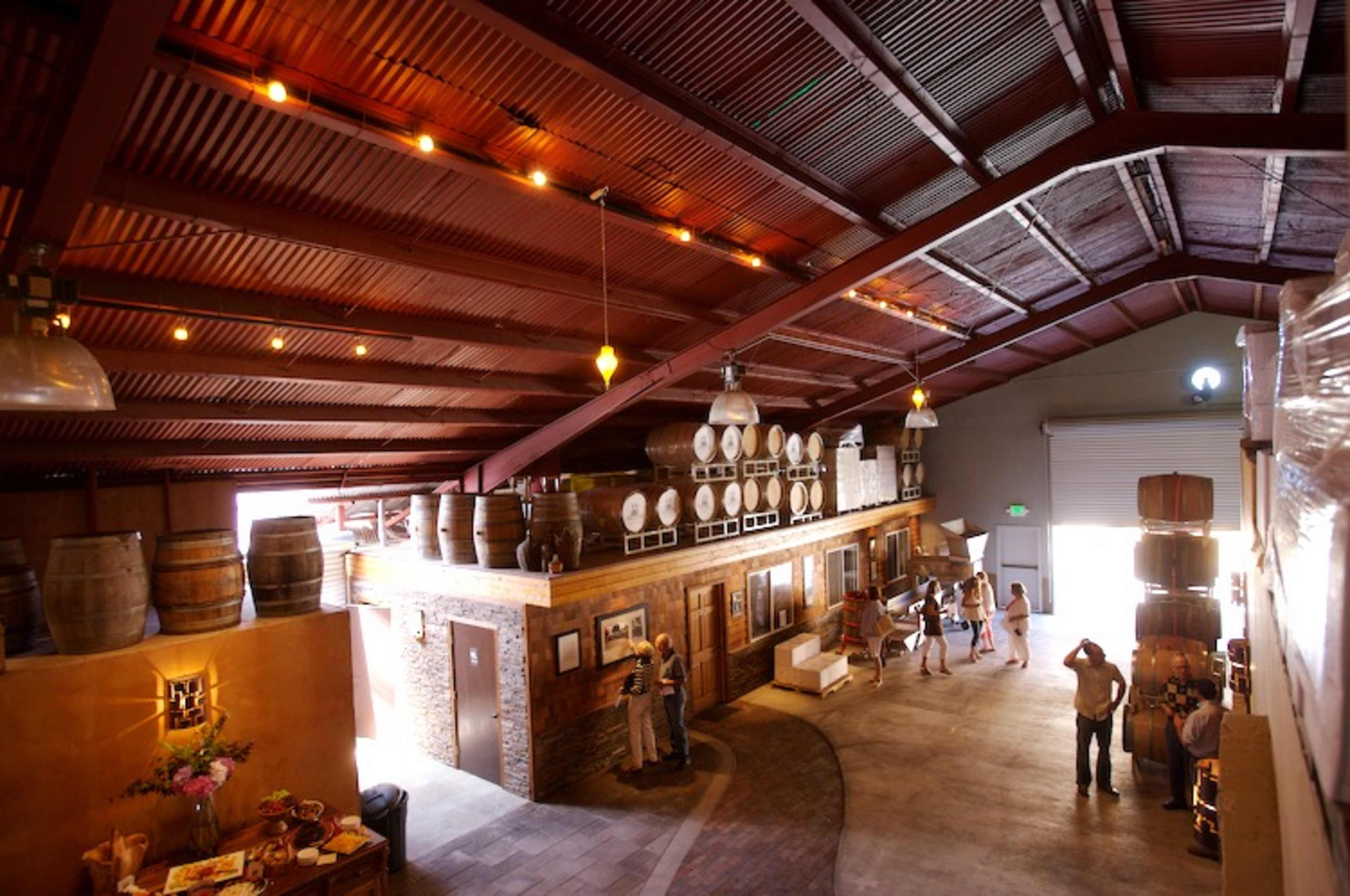 The image shows a spacious winery interior with high ceilings, wooden barrels, and several people engaged in conversation.