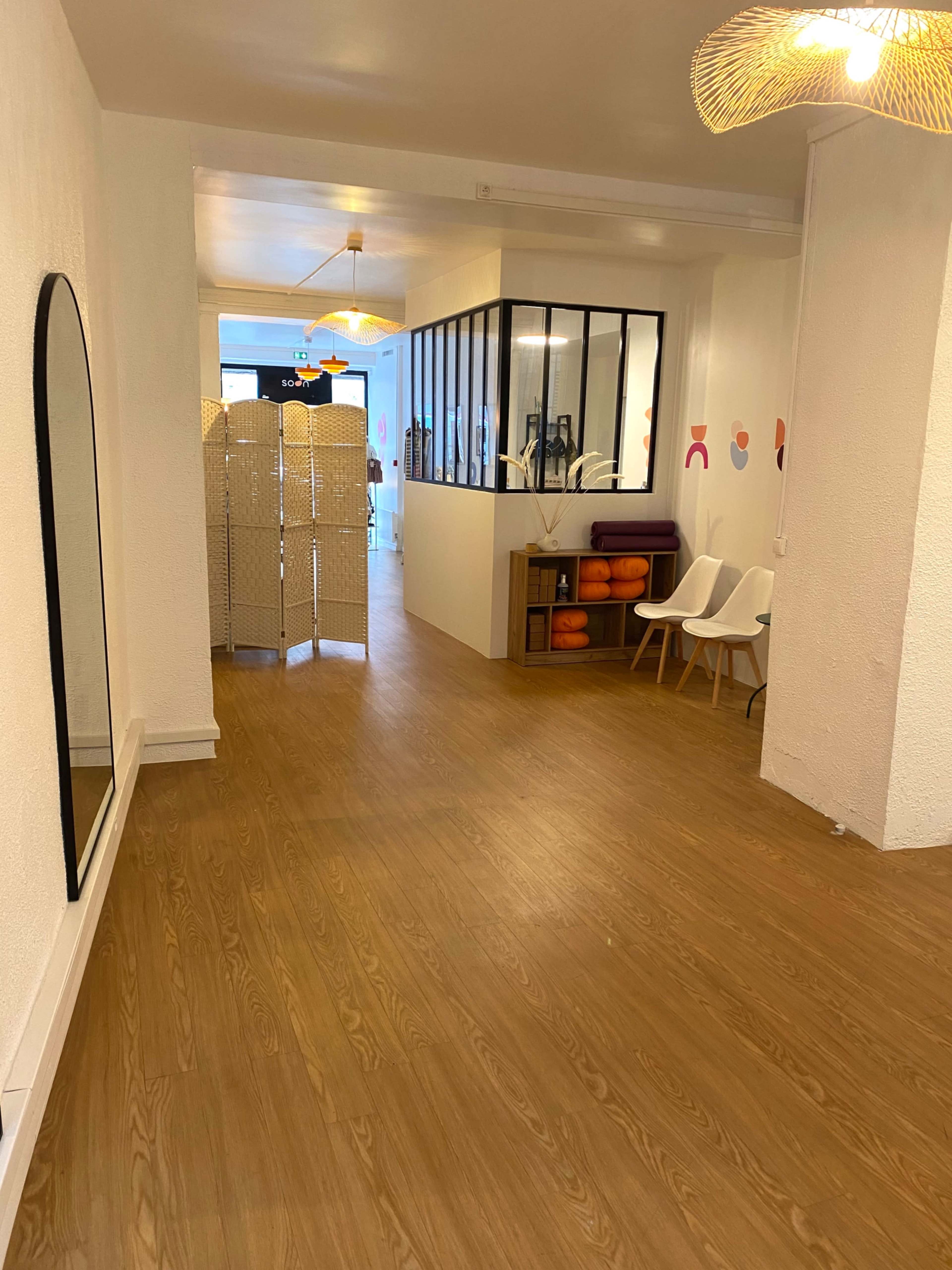 A well-lit hallway features wooden flooring, a large mirror on one wall, and a partition with a seating area visible in the background.